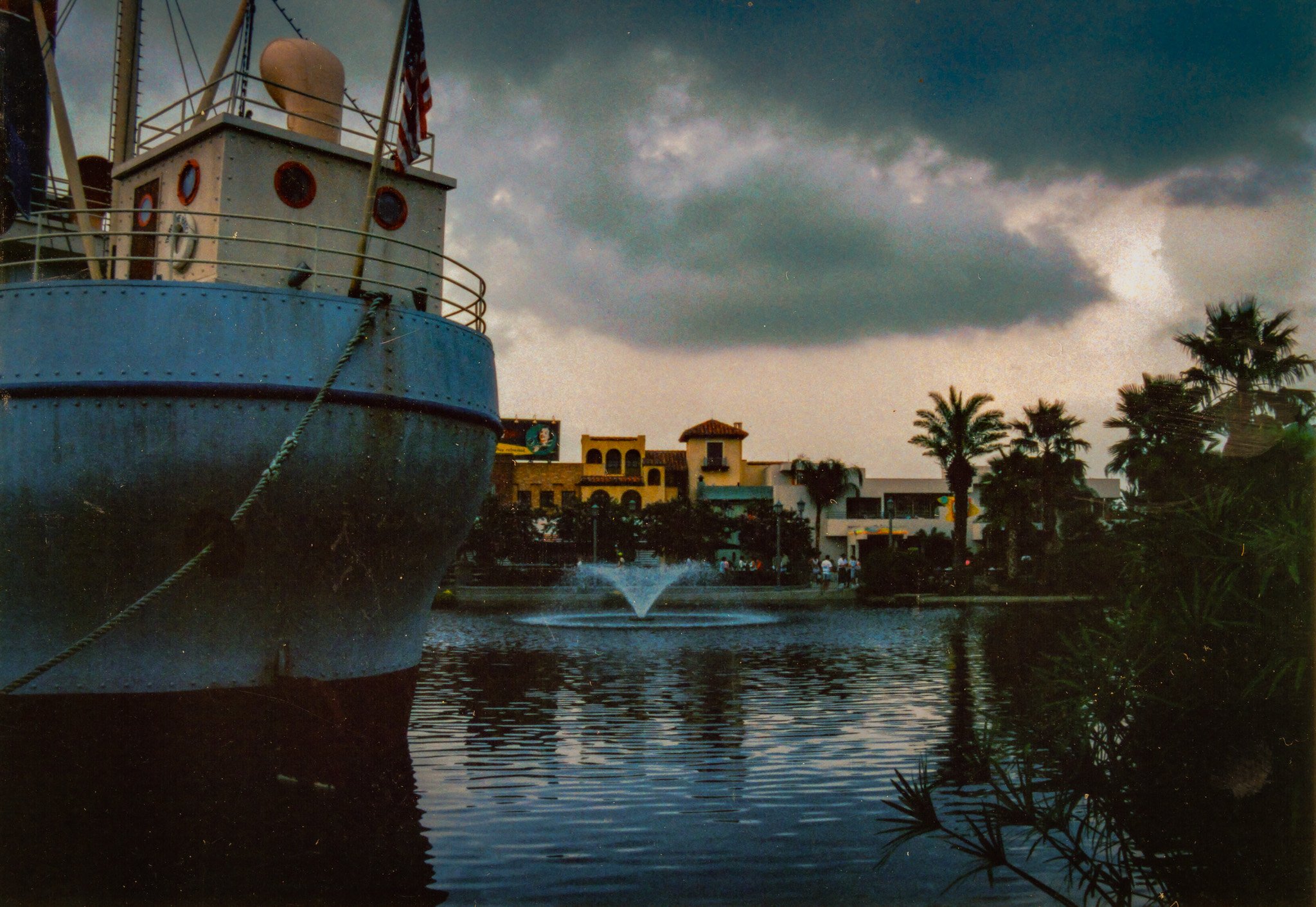 Image of a large boat in a waterway with splashing water at the front, surrounded by palm trees and buildings, with dark stormy clouds overhead during the late evening or early morning.