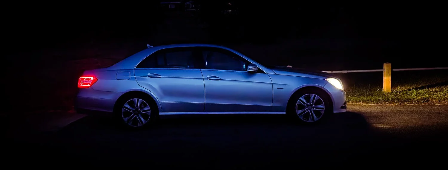 A silver sedan car with its headlights on, parked near a yellow post in a dark outdoor setting.