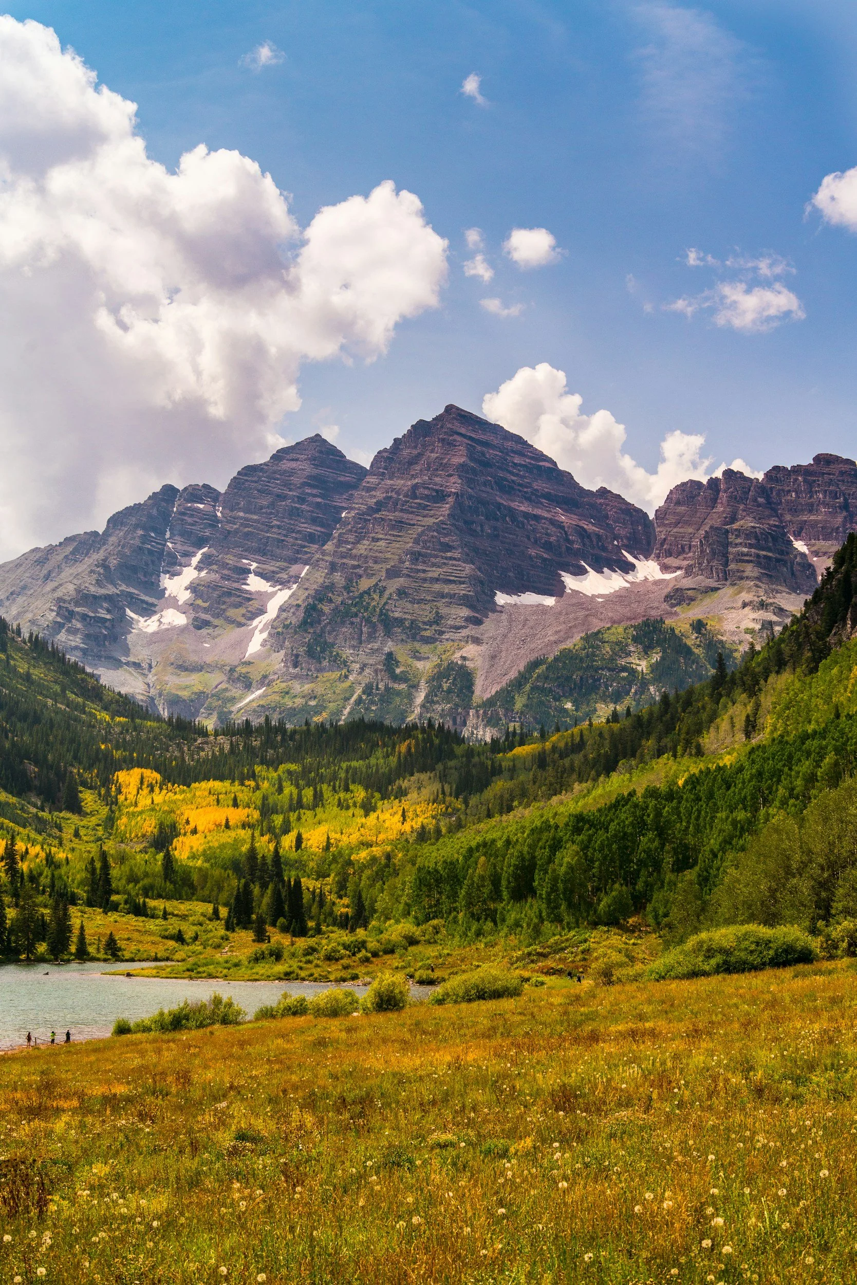 A scenic mountain landscape with rugged peaks, a lake in the foreground, and lush trees and grass in vibrant yellows and greens, under a partly cloudy blue sky.