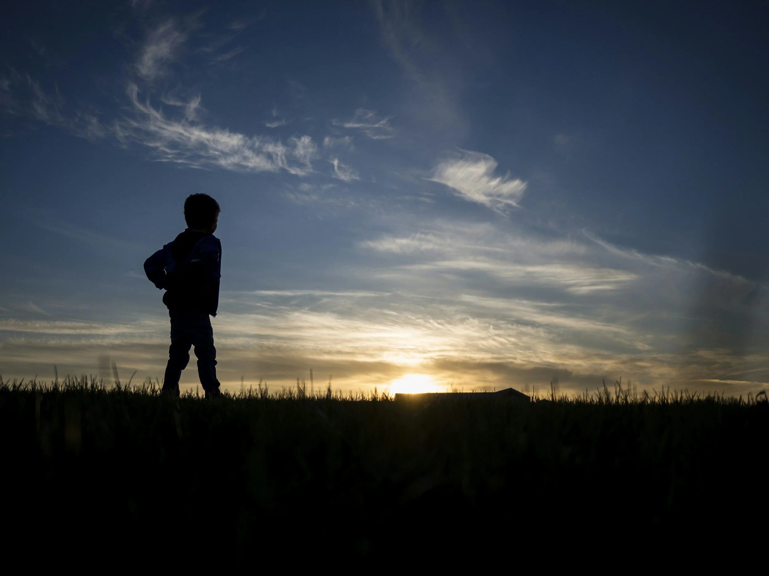 Silhouette of a person standing outdoors during sunset, with clouds in the sky and a grassy ground in the foreground.