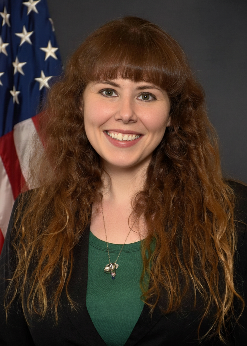 A woman with long, curly red hair smiling in front of a U.S. flag.