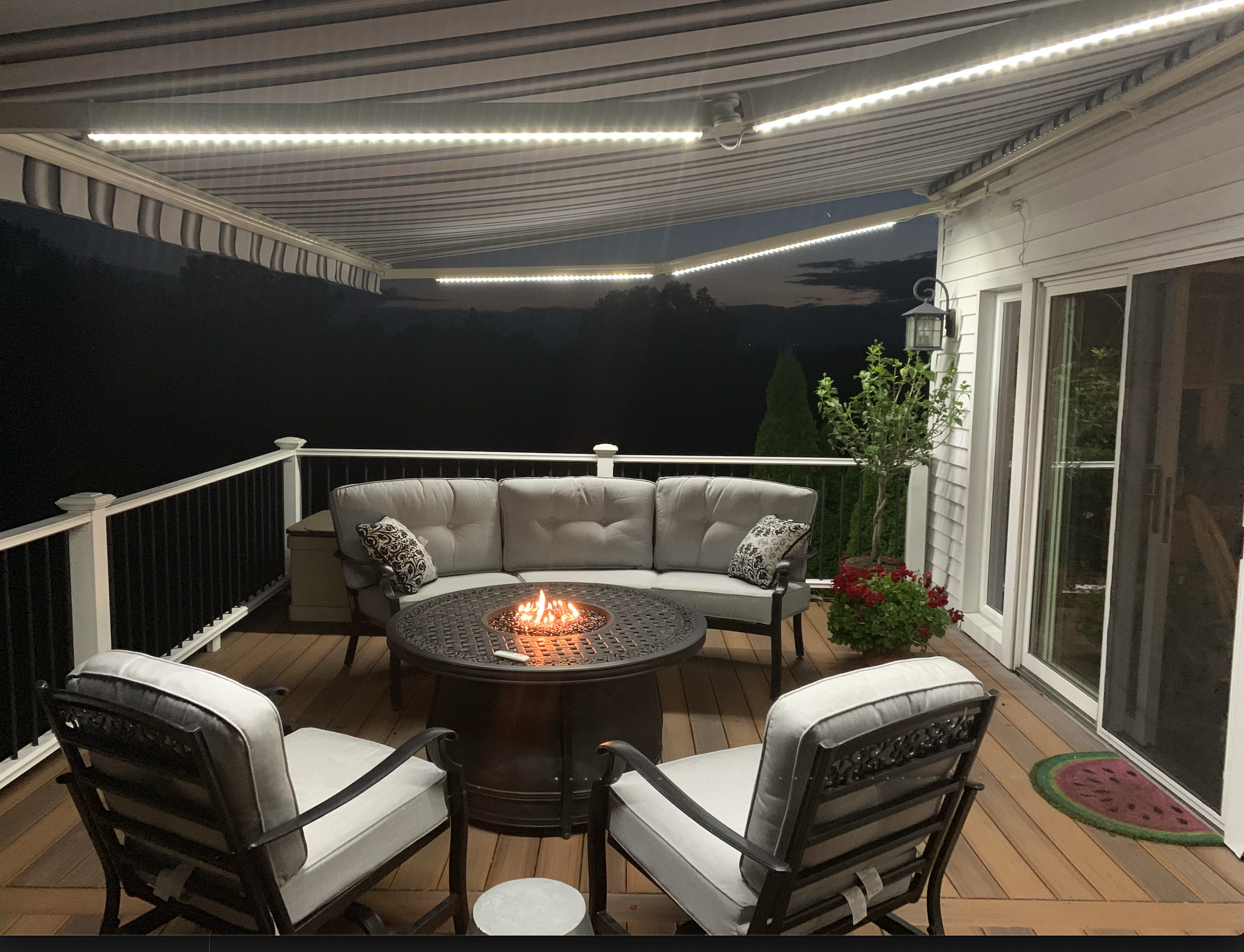A backyard patio with a firepit, surrounding chairs with cushions, a small side table, and potted plants. Overhead string lights illuminate the area, and the sky appears to be at dusk or night.
