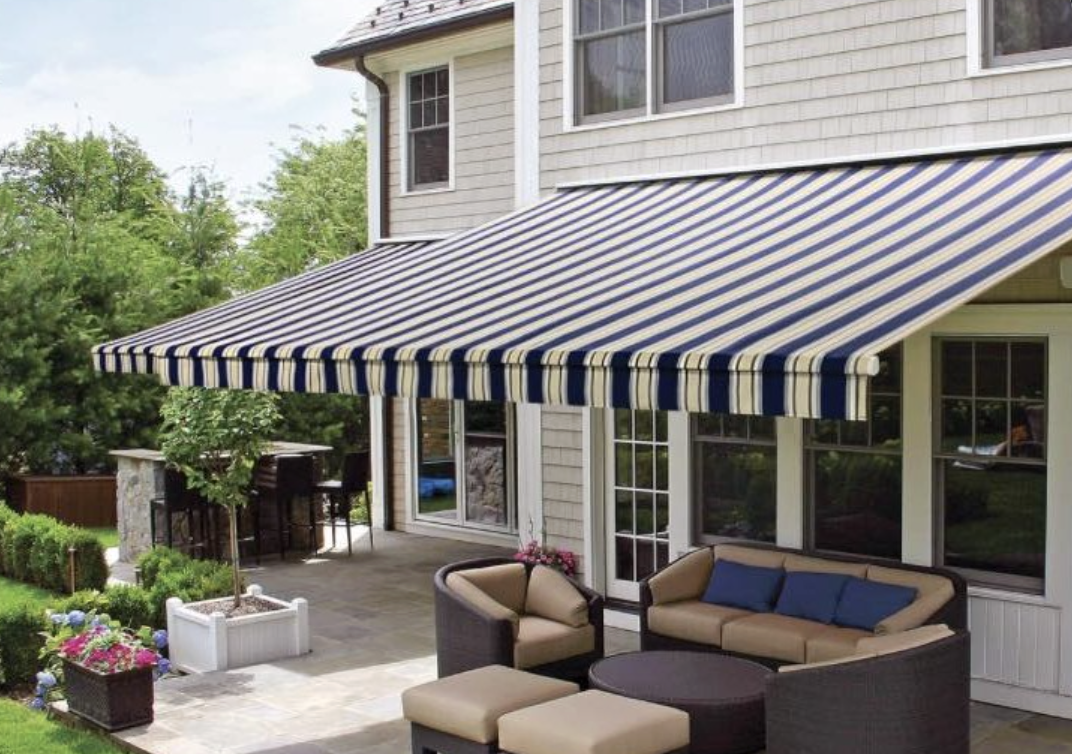 Backyard patio with outdoor seating, table, potted plants, small tree, and a striped blue and yellow awning attached to a house.