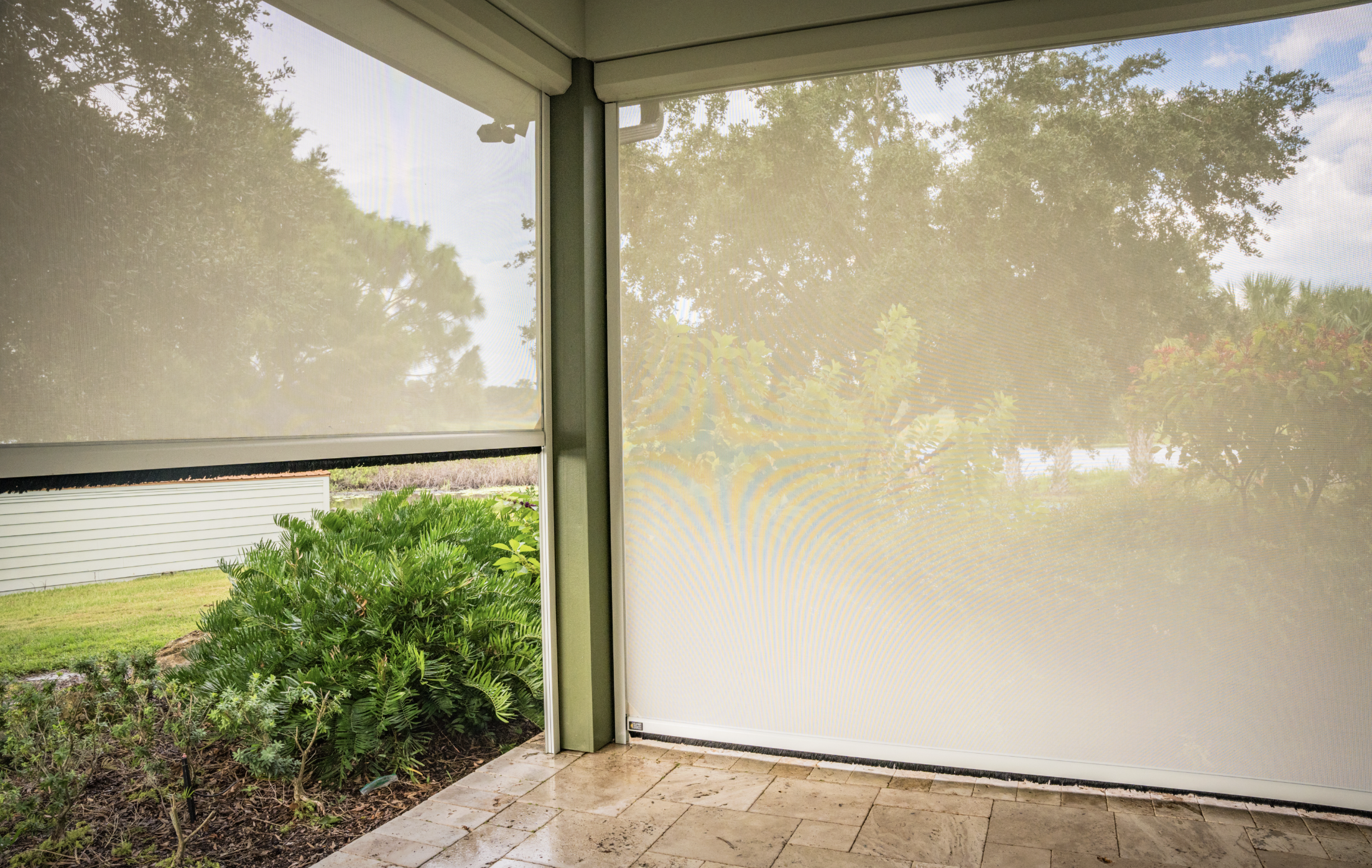 View from inside a screened porch looking out at a garden with lush green plants, trees, and a lawn, with a light-colored tiled floor and screened sliding doors.