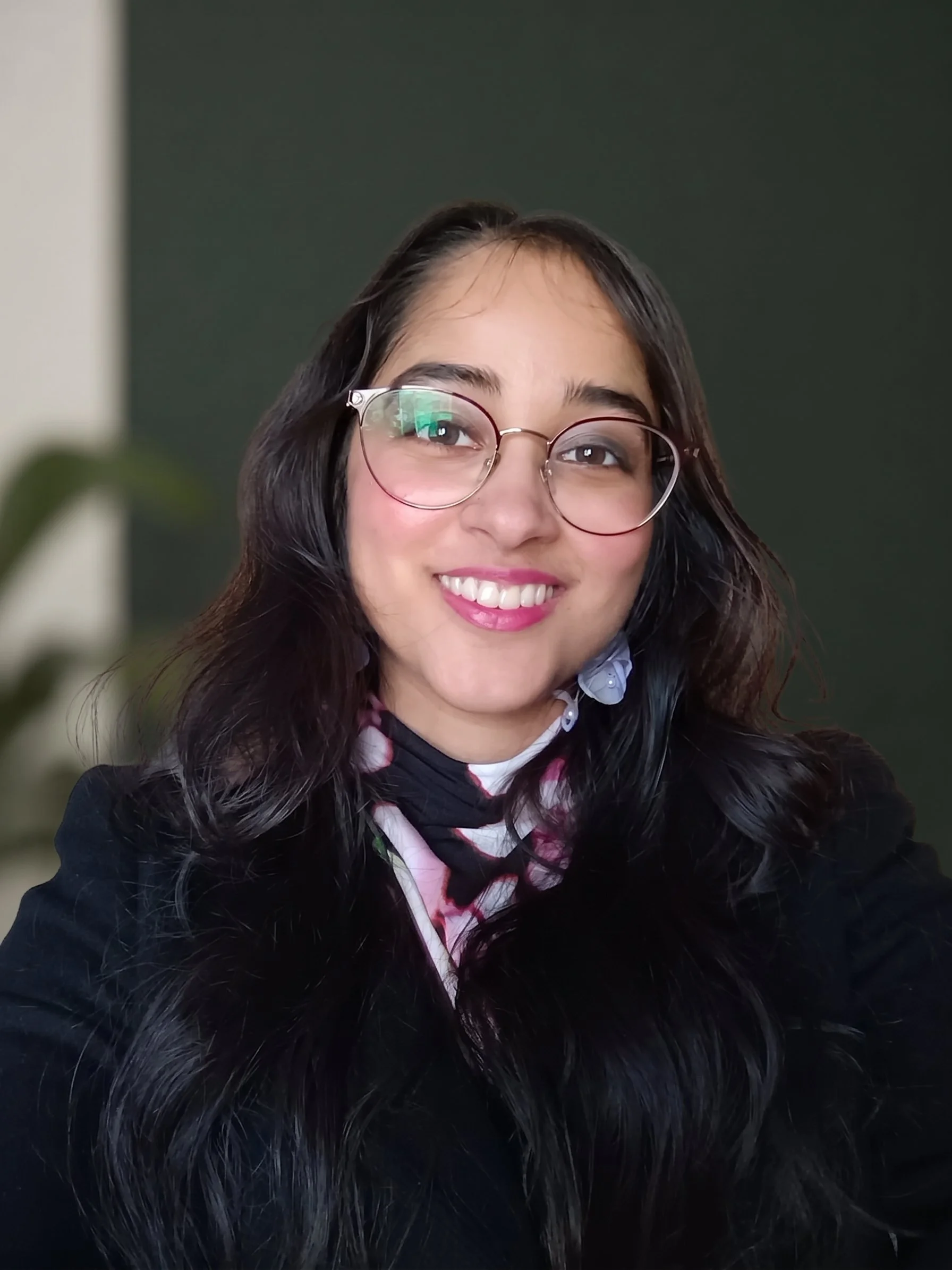 Smiling woman with glasses and long dark hair, wearing a black coat and patterned scarf, indoors against a dark background.