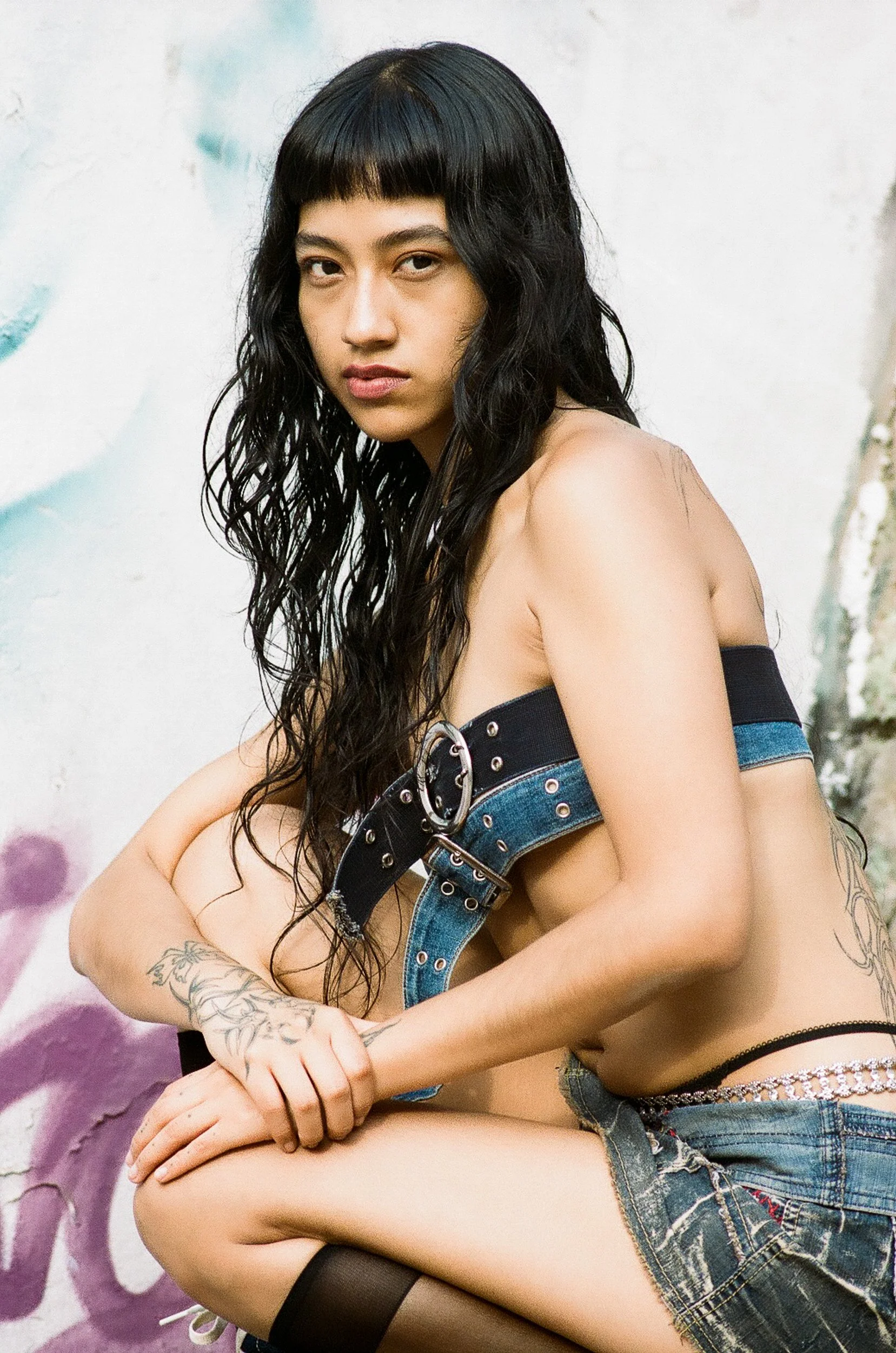 A young woman with long curly black hair, wearing a black strapless top, denim shorts, and thigh-high stockings, sitting against a graffiti wall background.
