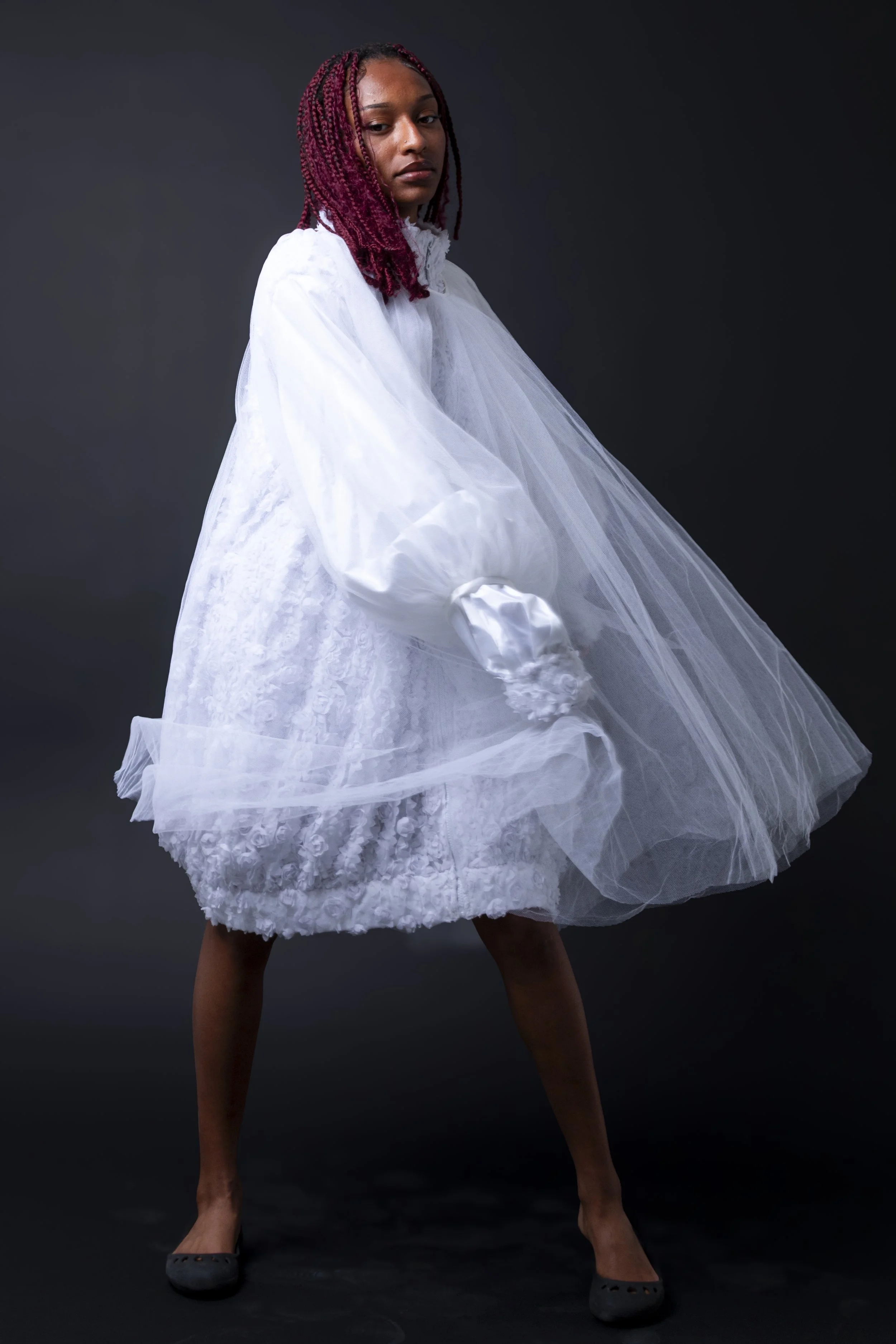 A woman with pink braided hair wearing a white layered dress with tulle and lace details, standing against a dark background.