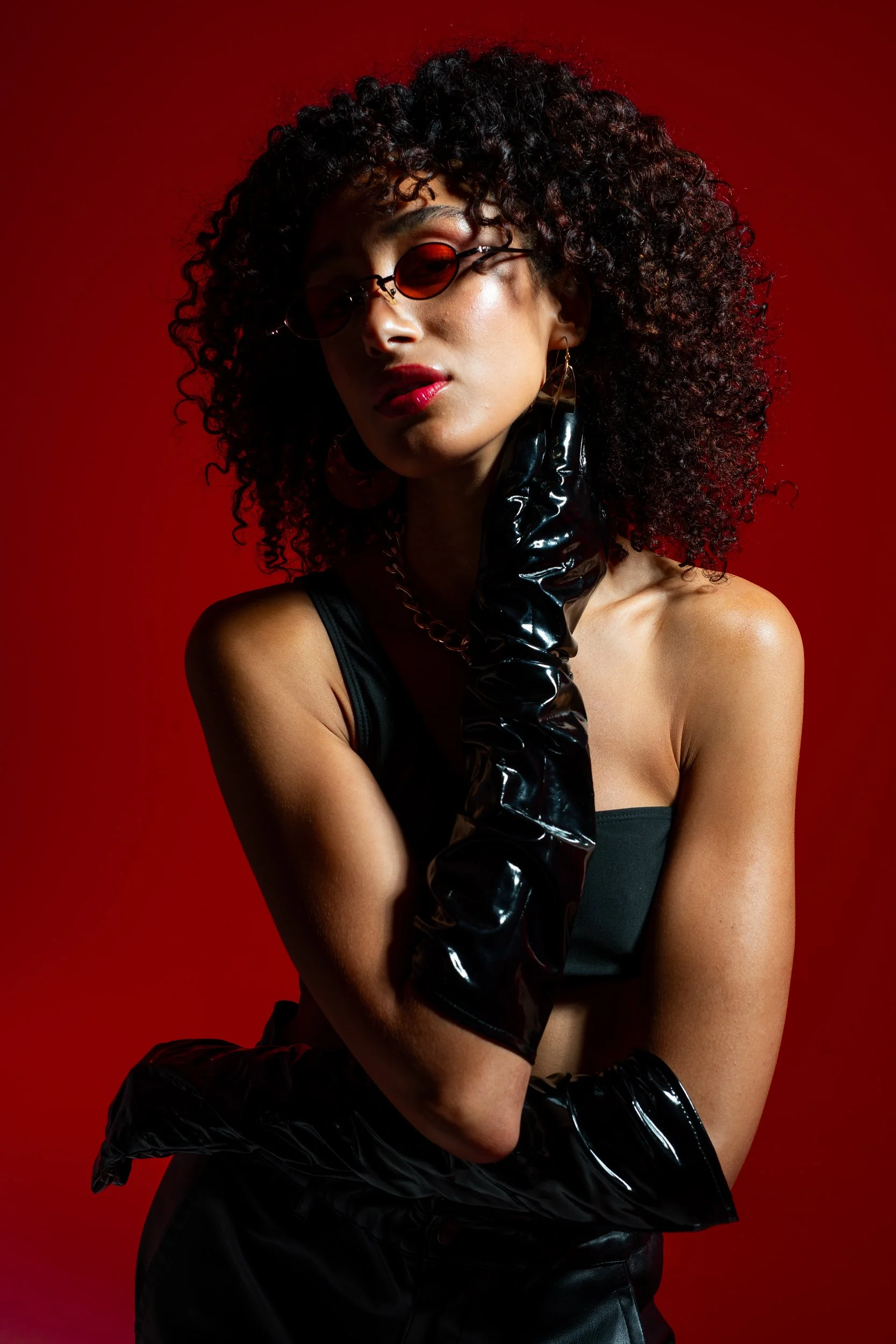 A woman with curly hair wearing red-tinted sunglasses, glossy black gloves, and black clothing poses against a red background.