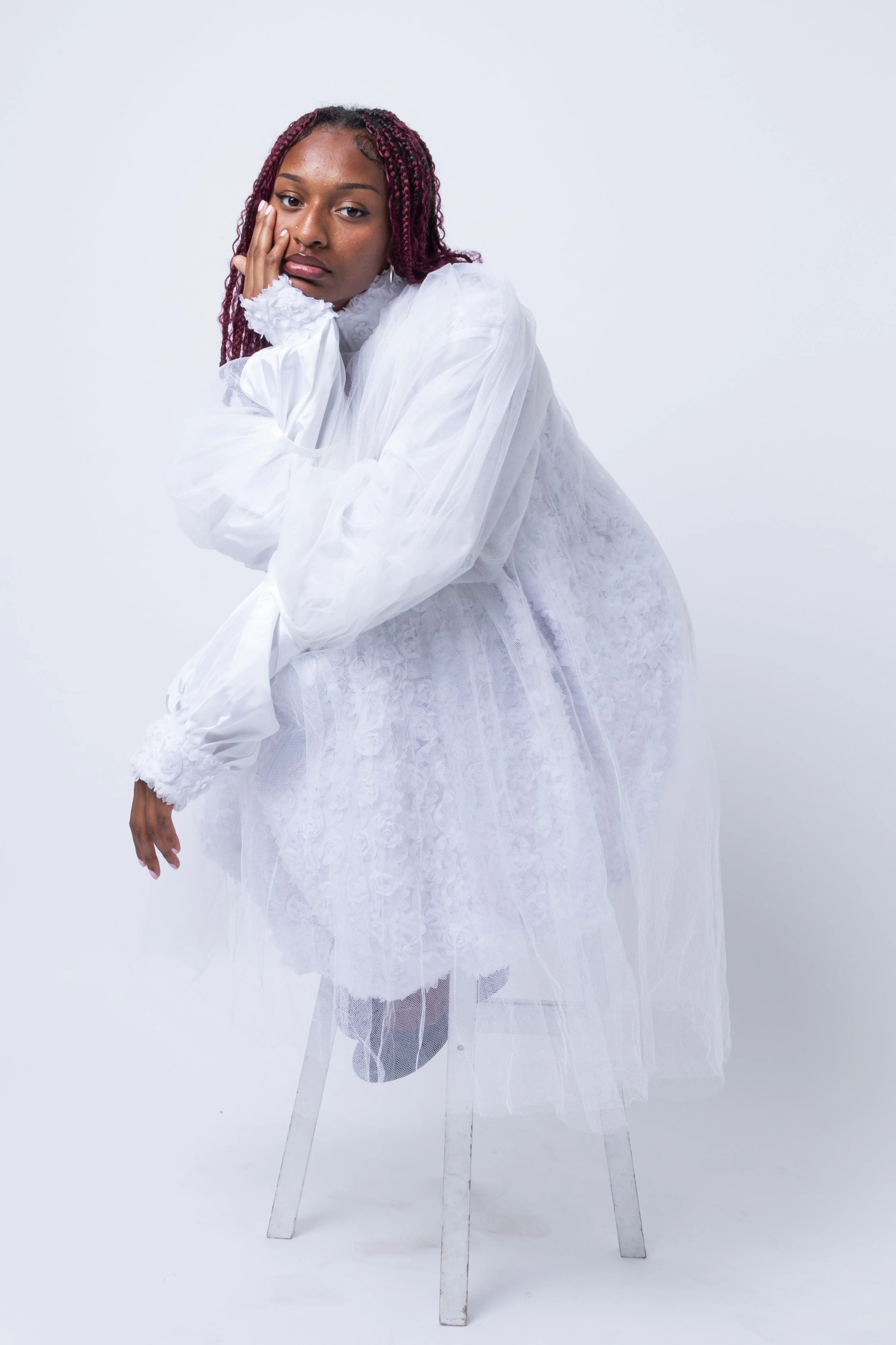 A woman with red and black twists wearing a white lace dress and standing against a white background.