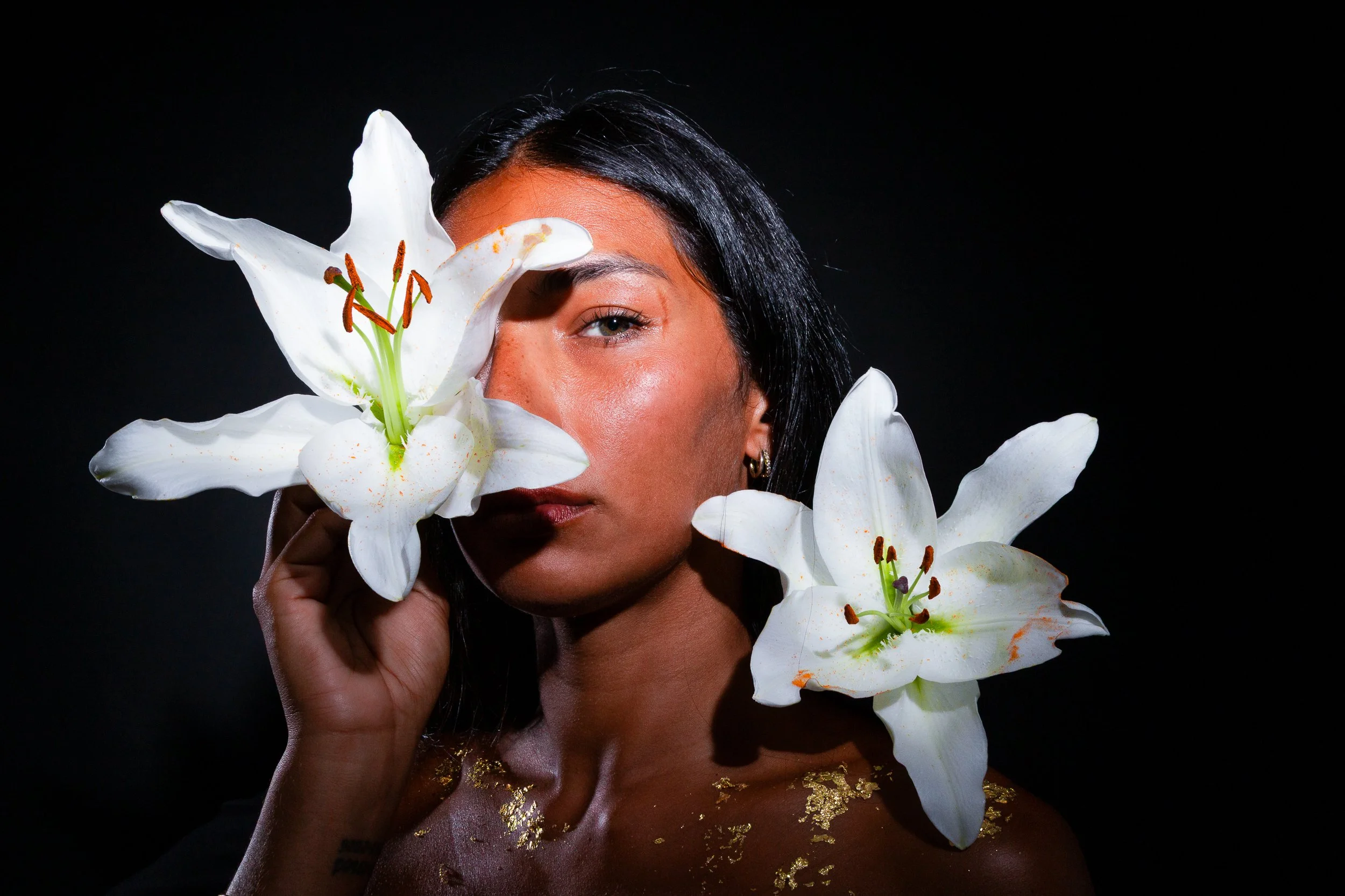 A woman with dark hair and tan skin holds white lilies with orange and black stamens close to her face against a black background.