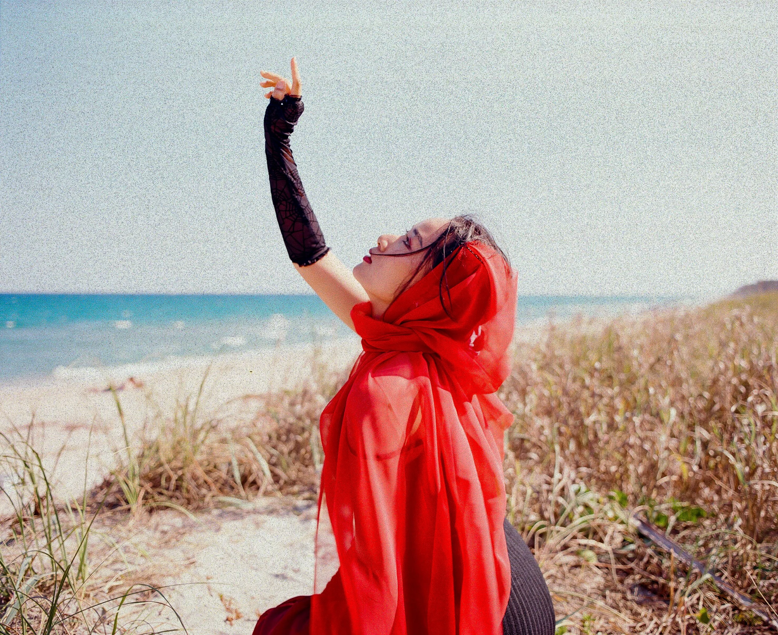 A woman standing outdoors at the beach, wearing a red cover-up and a black long-sleeve top, with her eyes closed and arms raised, facing the sky.