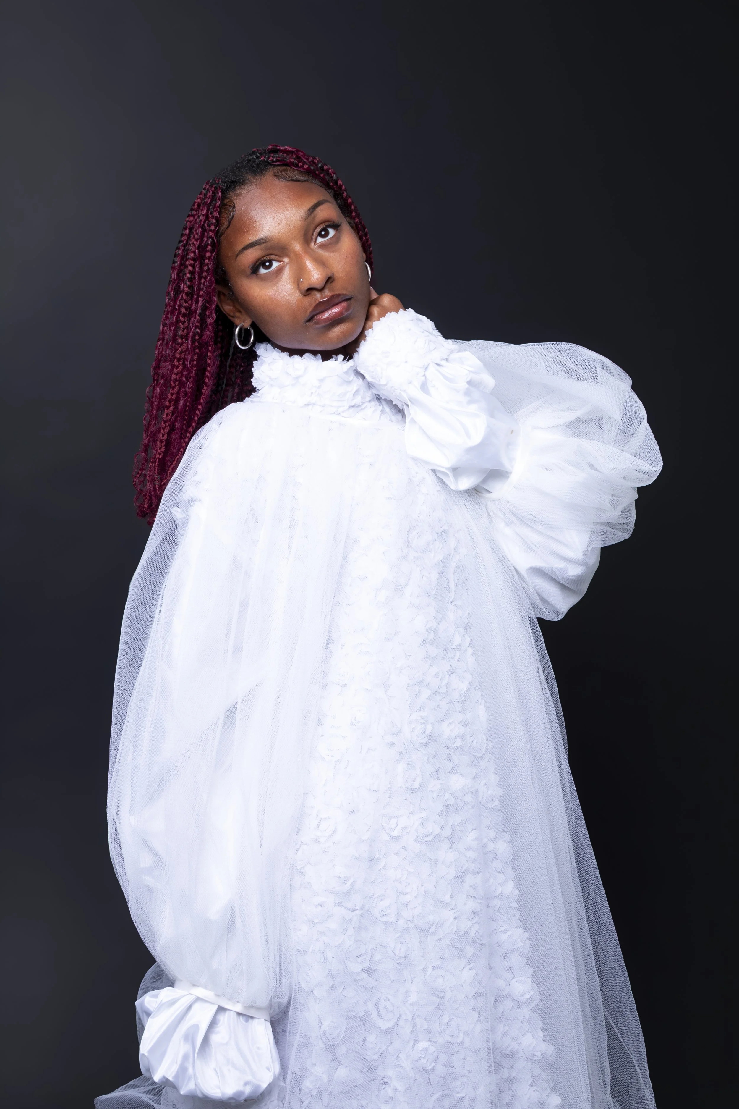 A young woman with red braids looking upwards wearing a white, textured dress with puffed sleeves and a high collar, standing against a dark background.
