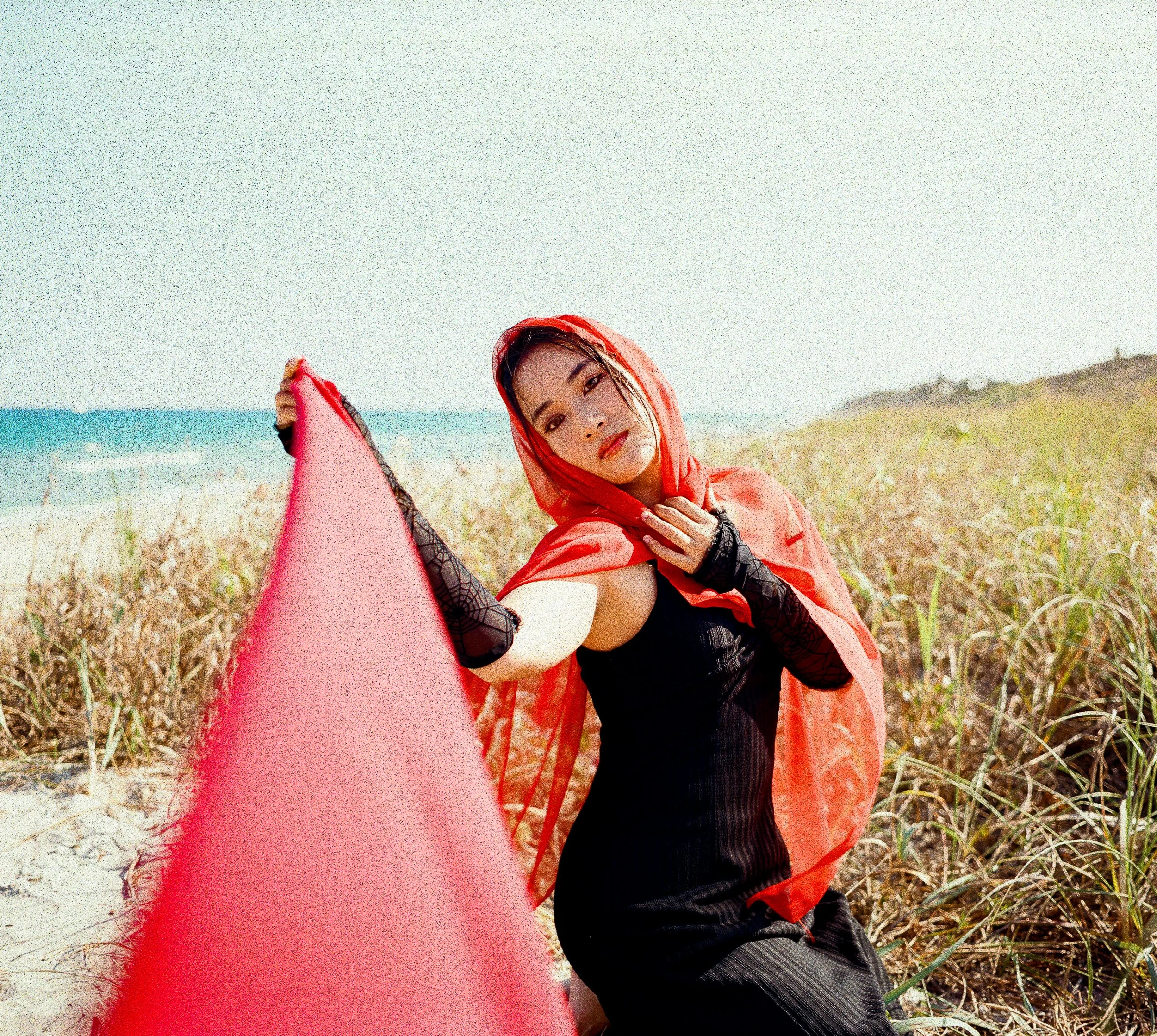 A woman with long black hair and makeup, wearing a red hoodie, holding a pink surfboard on a grassy beach landscape with the ocean in the background.