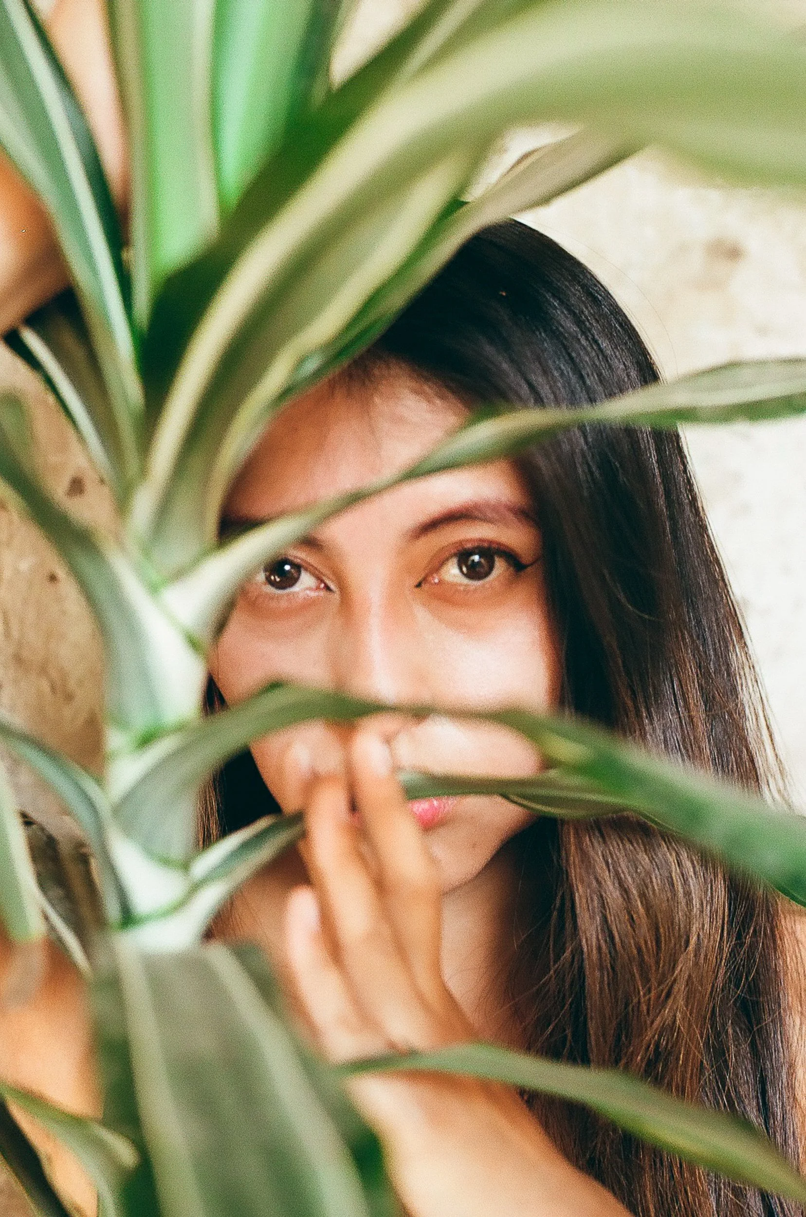 A woman with dark hair peeking through green plant leaves, her eyes and part of her face visible.