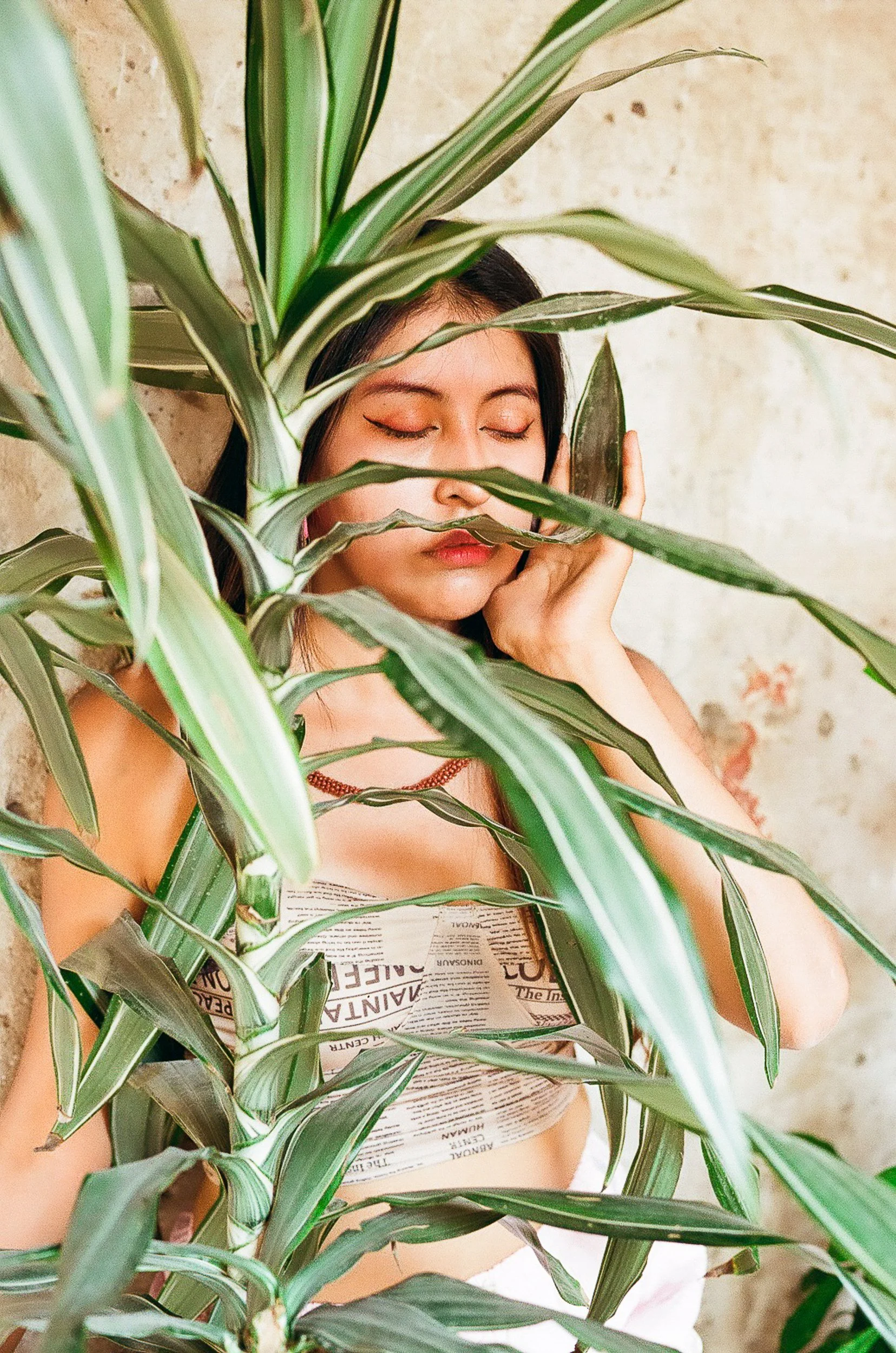 A woman with closed eyes and makeup, holding her face with one hand, standing behind green leafy plants and a wall with some paint peeling.