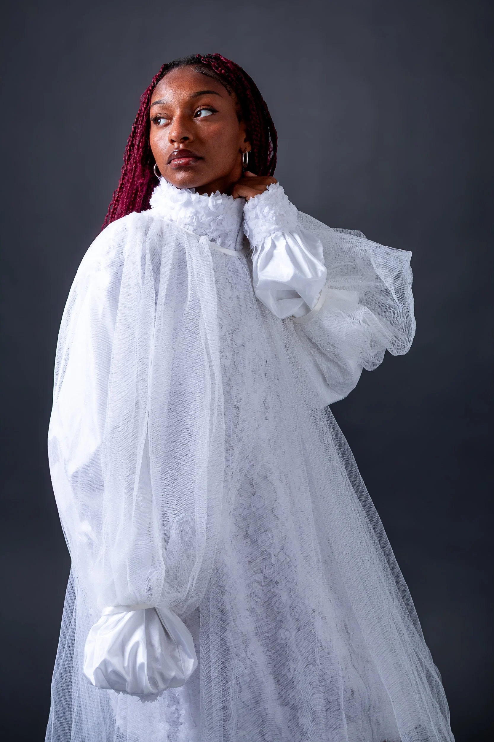 A woman with red braided hair wearing a white dress with puffed sleeves and ruffled collar, standing against a gray background.