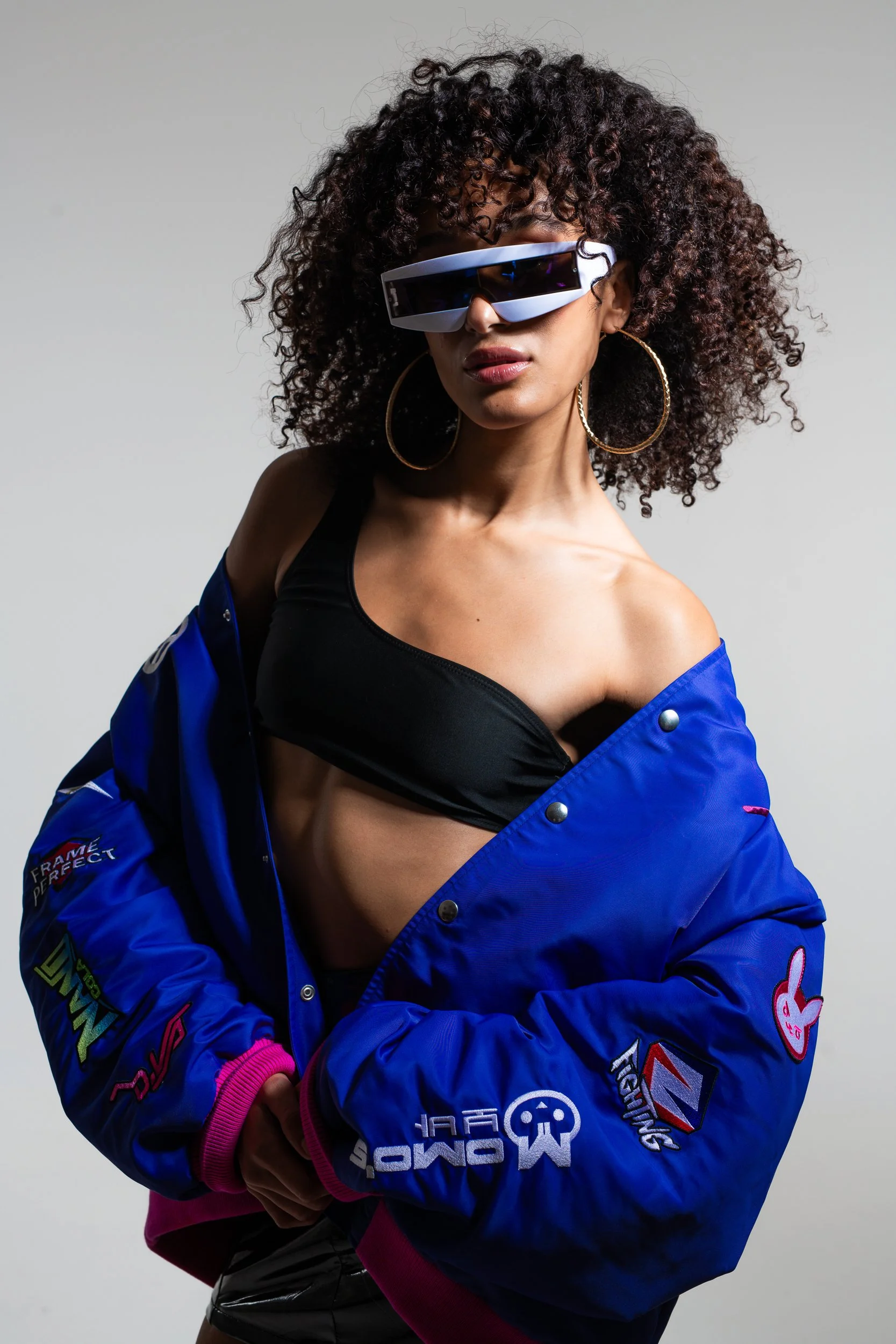 A woman with curly hair wearing large futuristic sunglasses, earrings, a black top, and a blue racing jacket with various patches and logos, posing against a neutral background.
