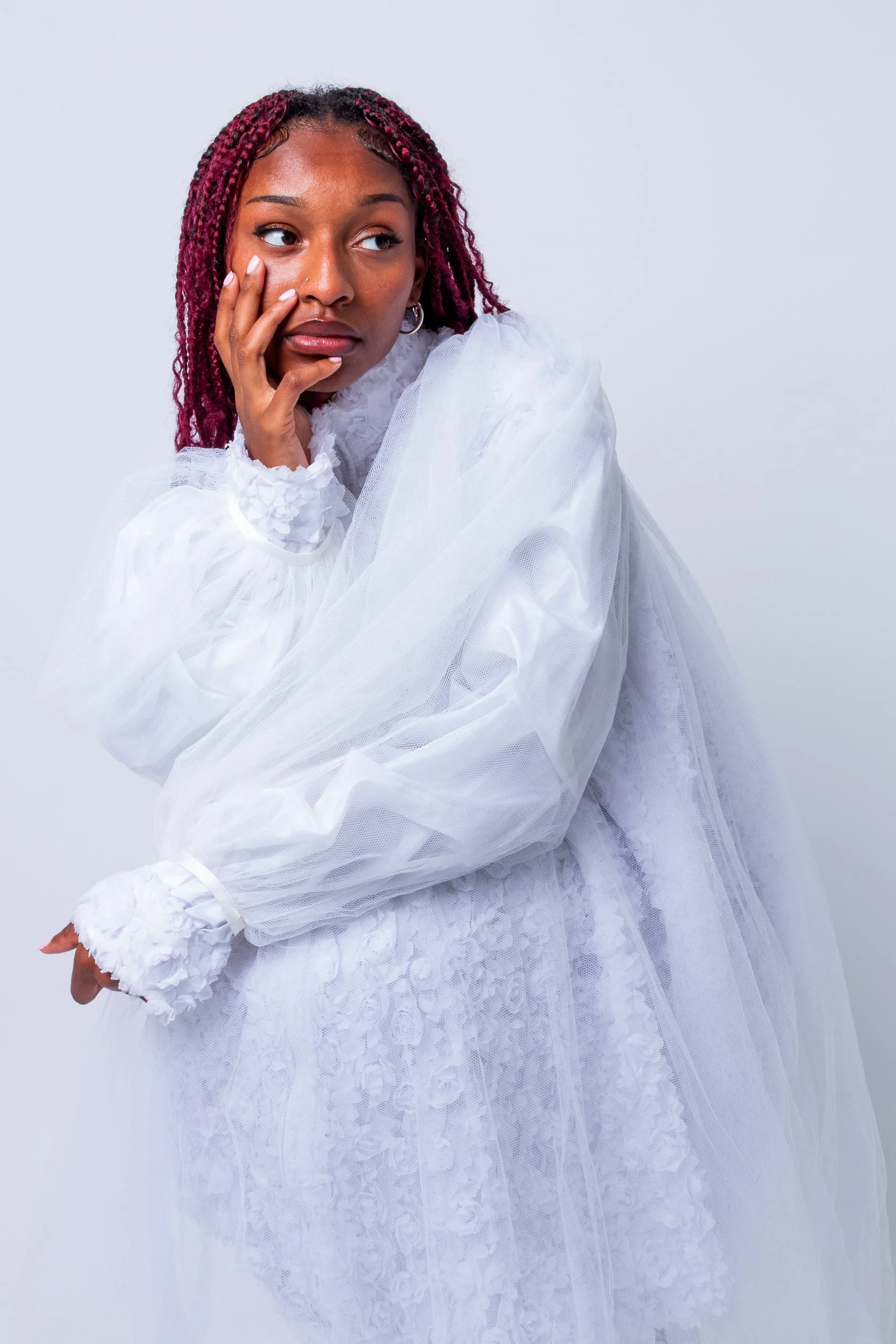 A woman with pink braids wearing a white dress with lace details, posing against a light background.