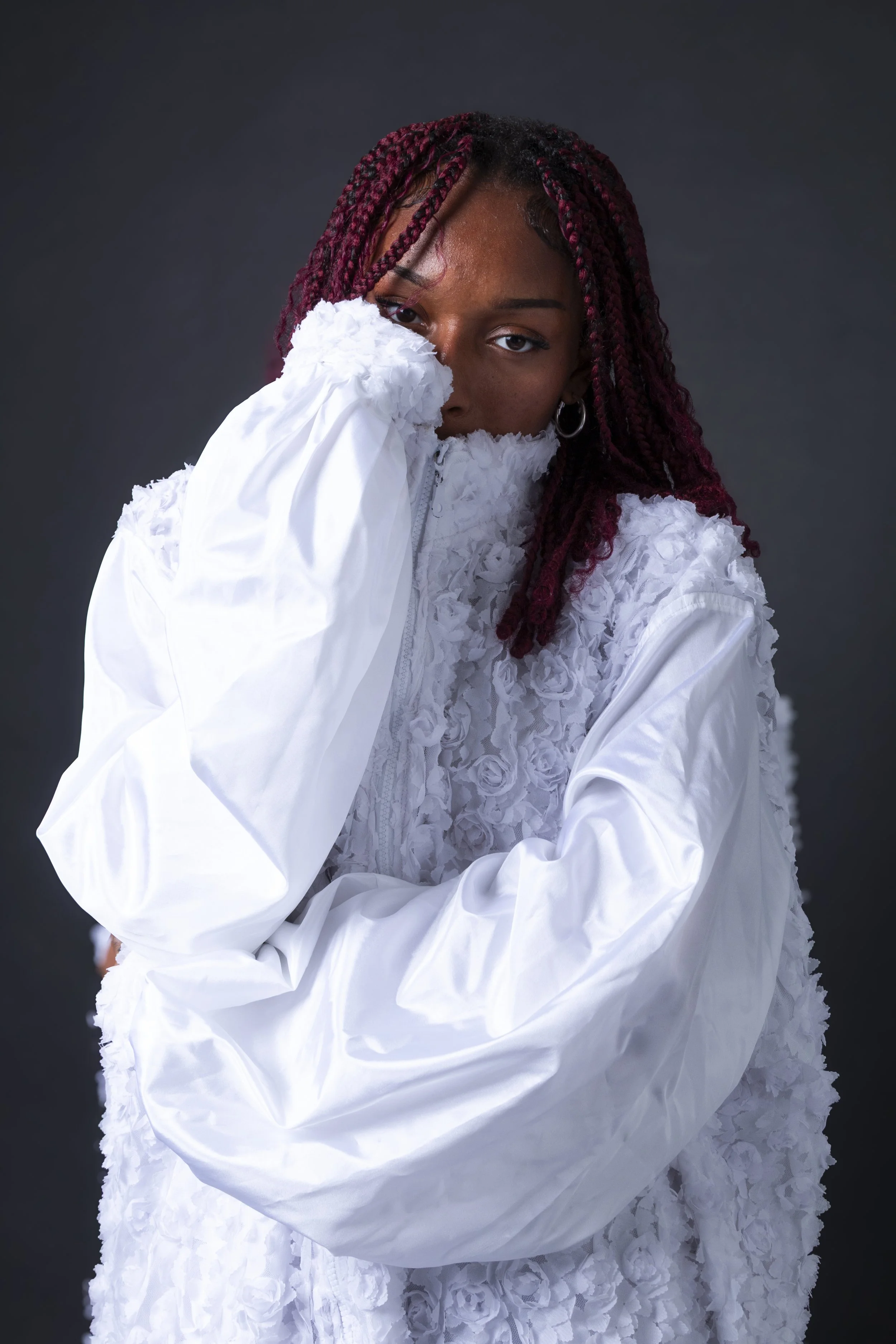 A woman with red braided hair covering part of her face, wearing a white textured jacket, against a dark background.