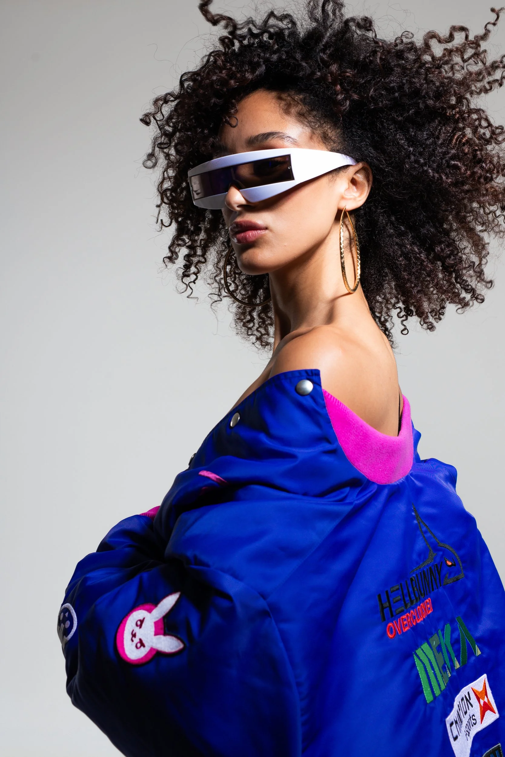 A woman with curly hair wearing futuristic sunglasses and colorful clothing with patches and text, posing against a plain gray background.