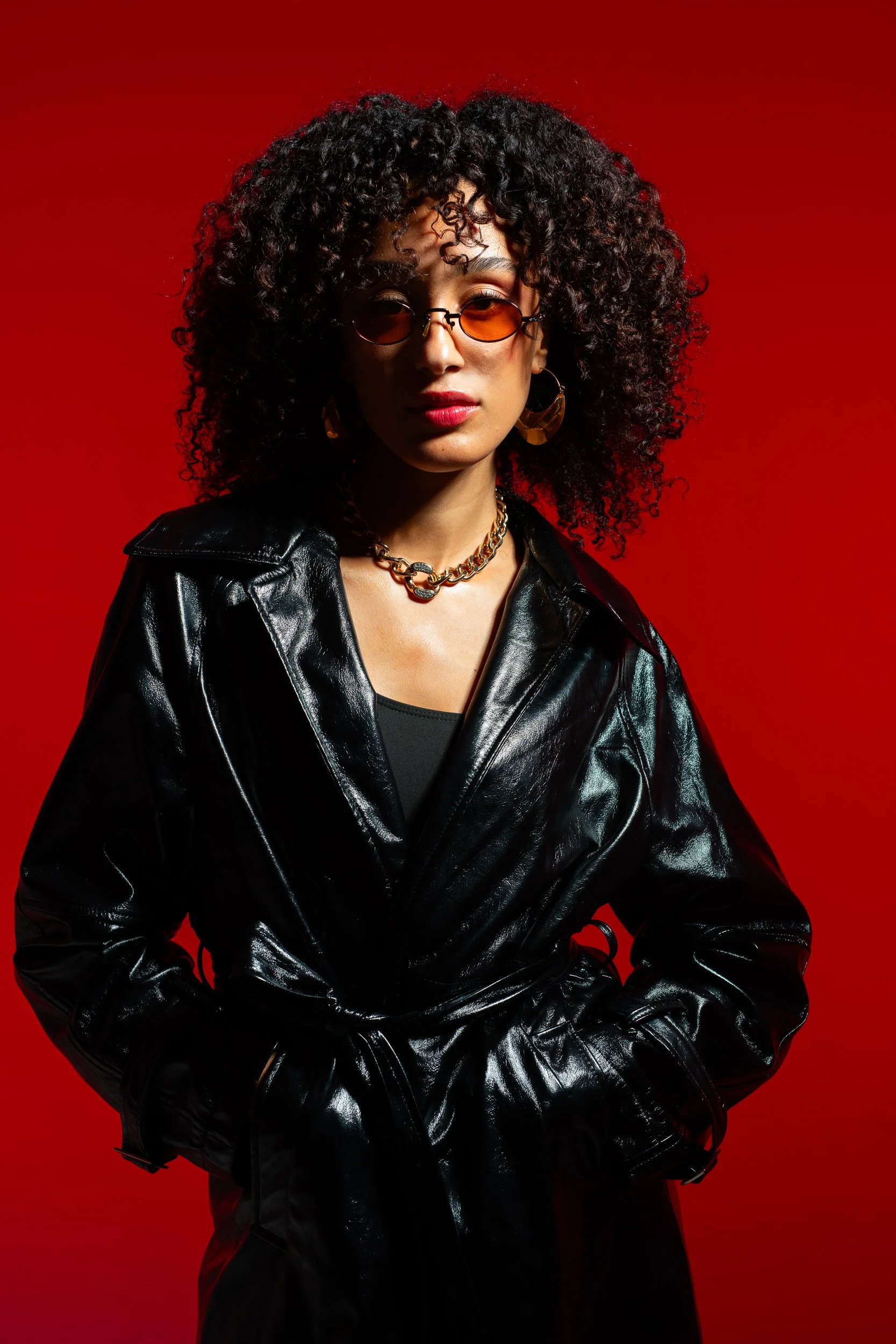 A woman with curly hair wearing orange-tinted sunglasses, gold hoop earrings, a gold chain necklace, a black shirt, and a black leather jacket against a red background.