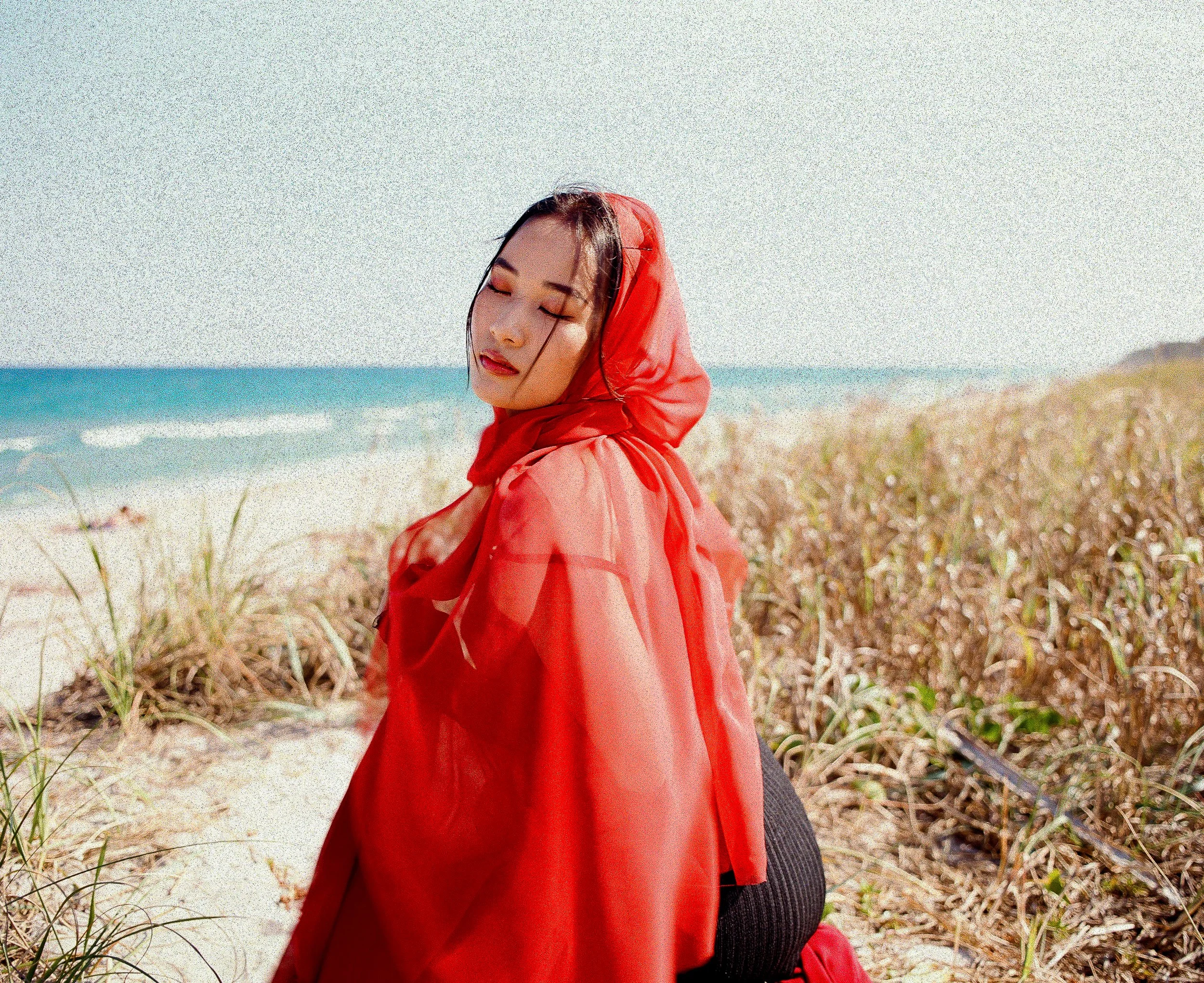 A woman with closed eyes wearing a red hooded raincoat at the beach with grass and sand in the foreground and the ocean in the background.