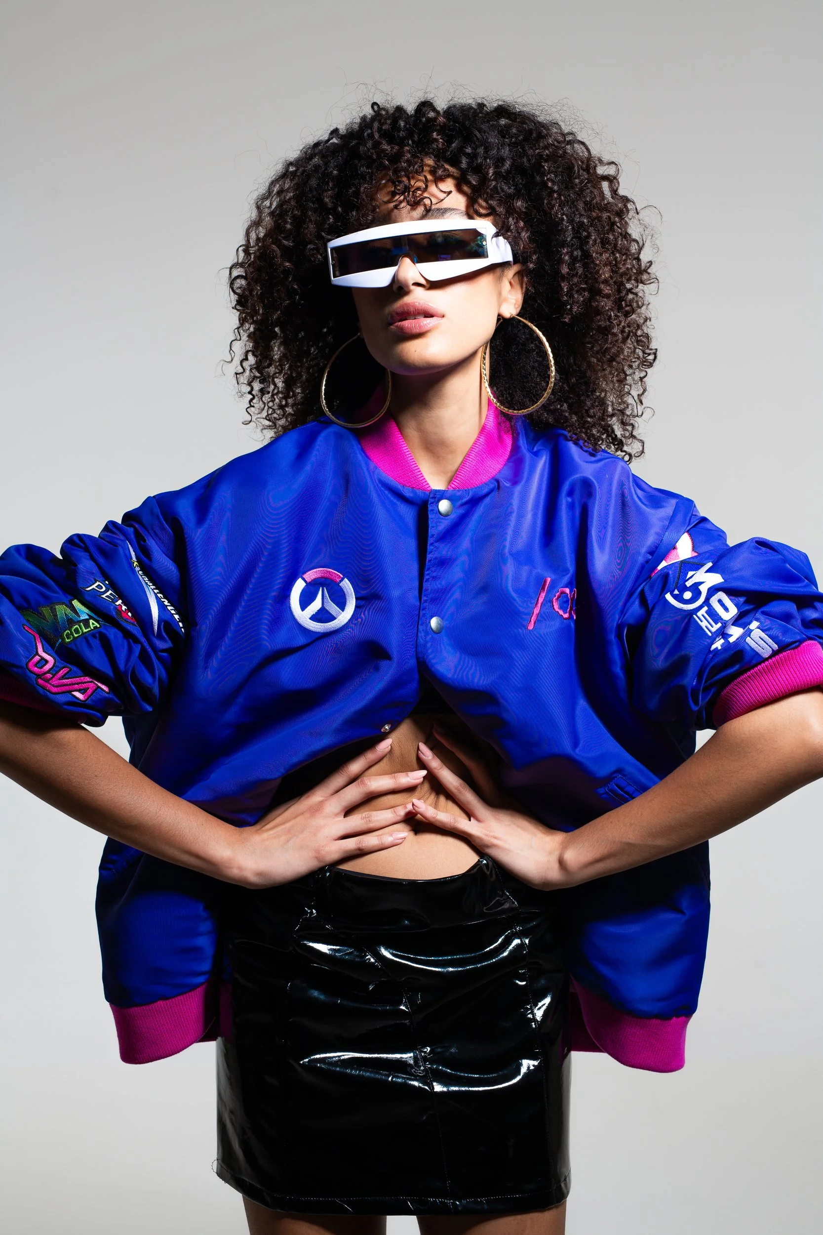 Woman with curly hair wearing futuristic sunglasses, a blue jacket with patches and logos, a black shiny skirt, pink socks, and large hoop earrings, standing against a plain gray background.