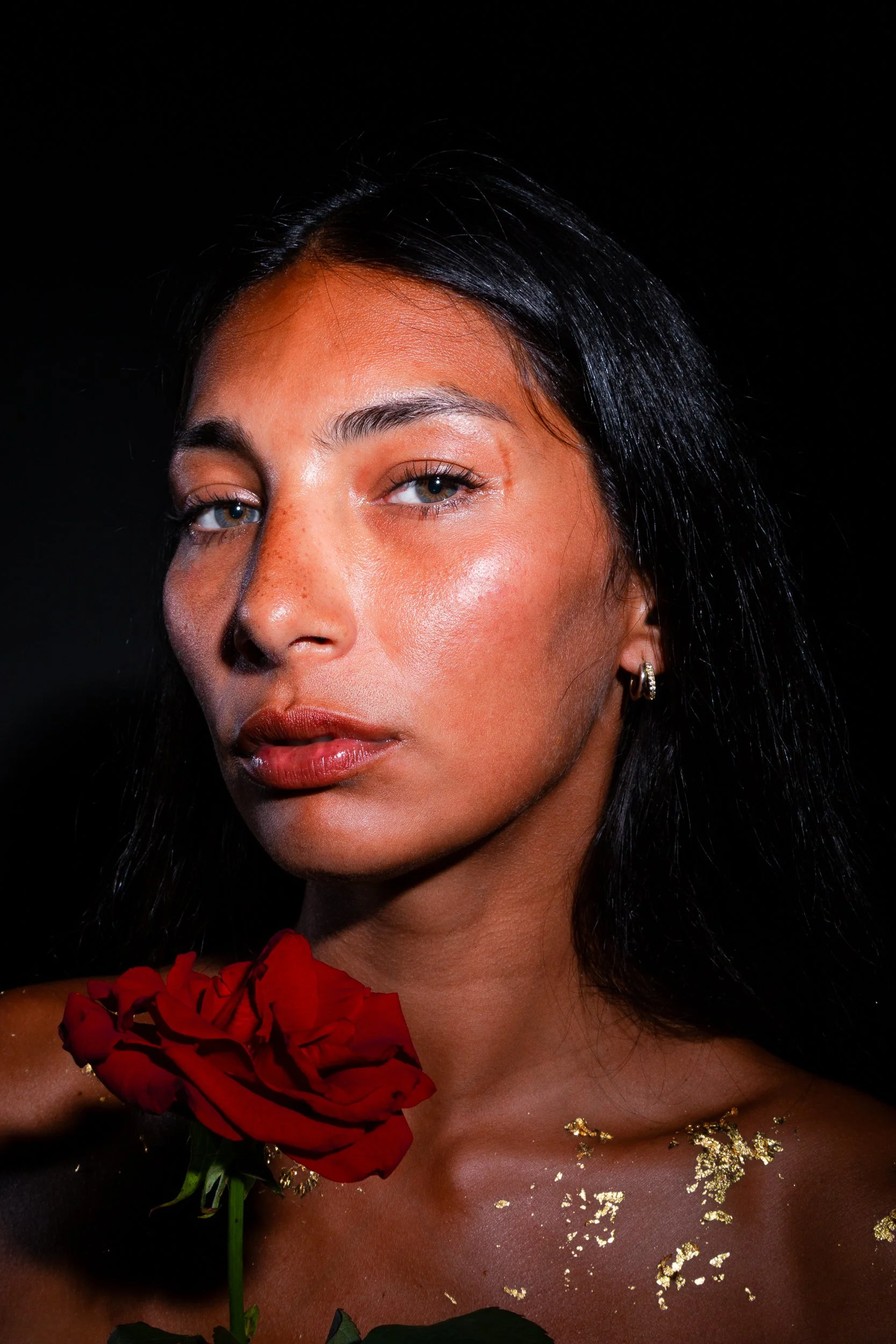 Close-up of a woman with dark hair, blue eyes, and light makeup, holding a red rose close to her face, with gold leaf accents on her skin and a black background.