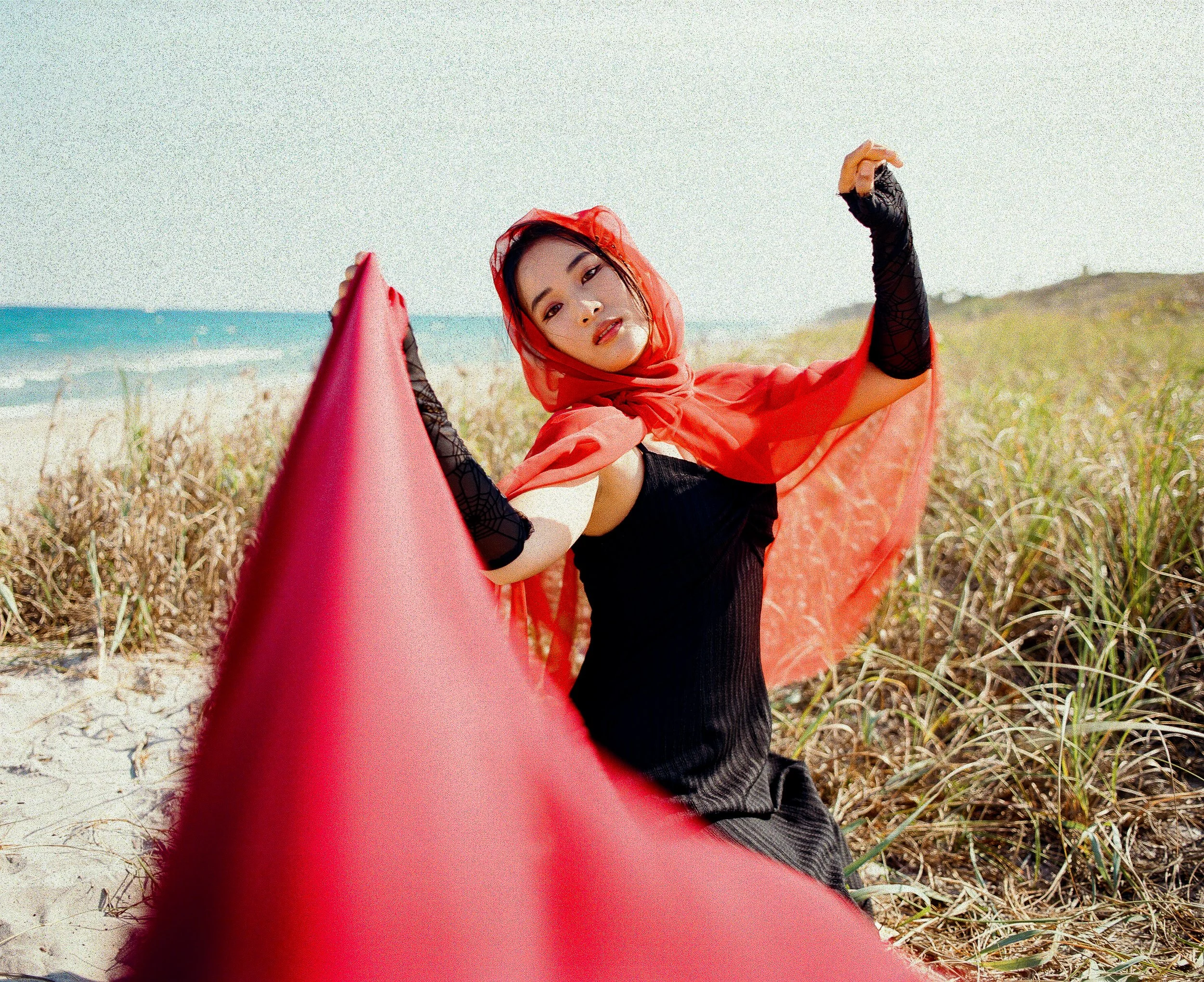 A woman wearing a red veil outdoors with beach and grassy dunes in the background, taking a selfie on her phone.