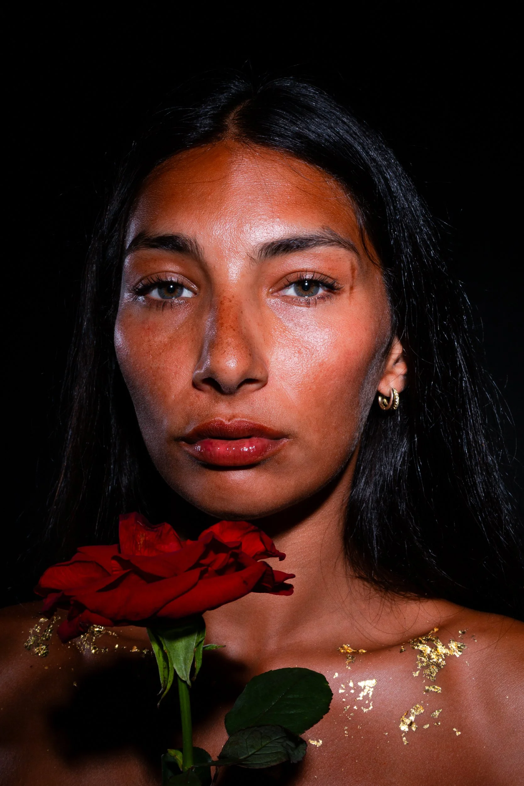 A woman with dark hair and light skin holding a red rose, decorated with gold leaf accents on her chest and shoulders, against a black background.