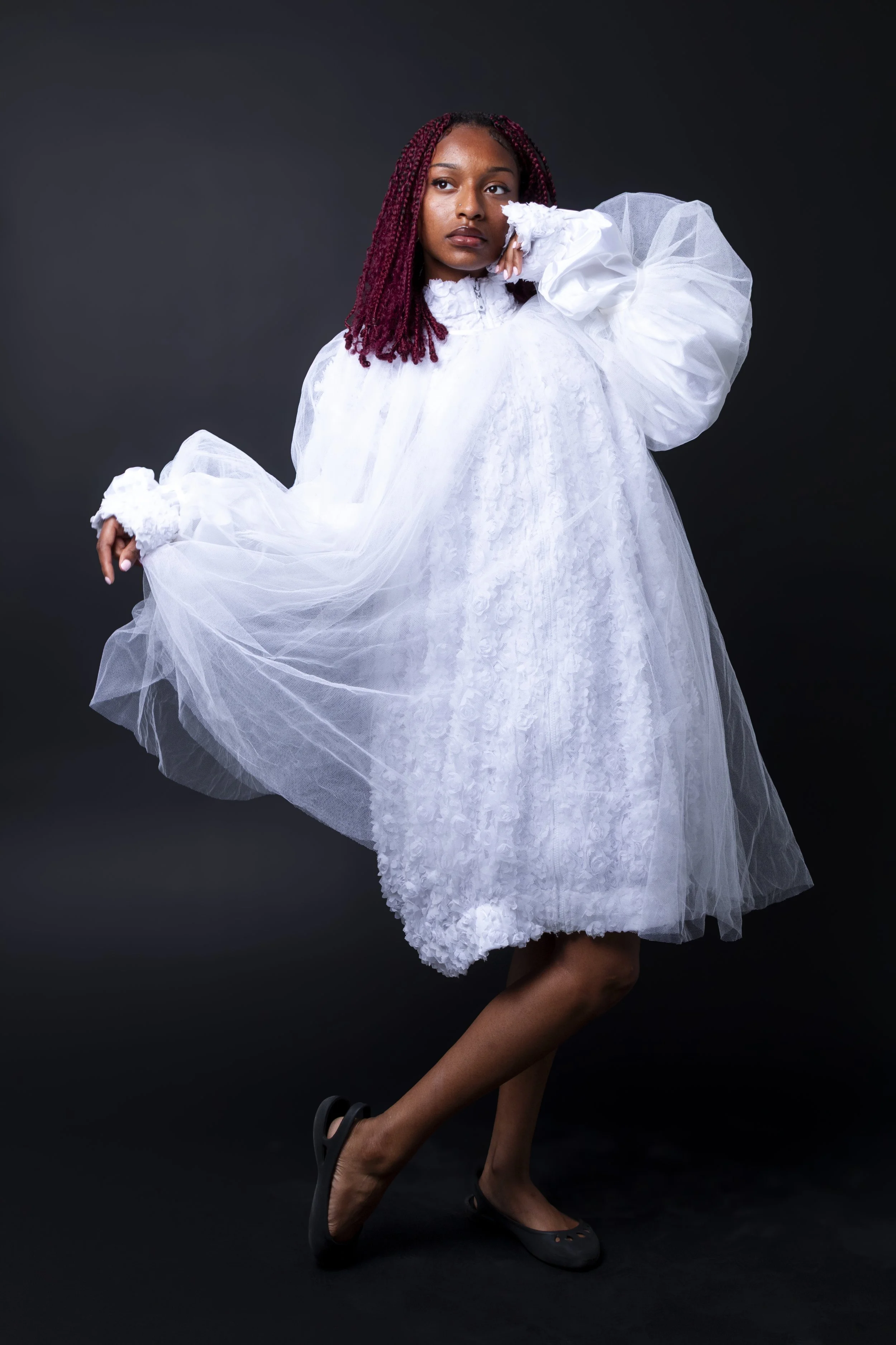 A woman with reddish braided hair poses in a white, voluminous dress with sheer tulle fabric, standing against a dark background.