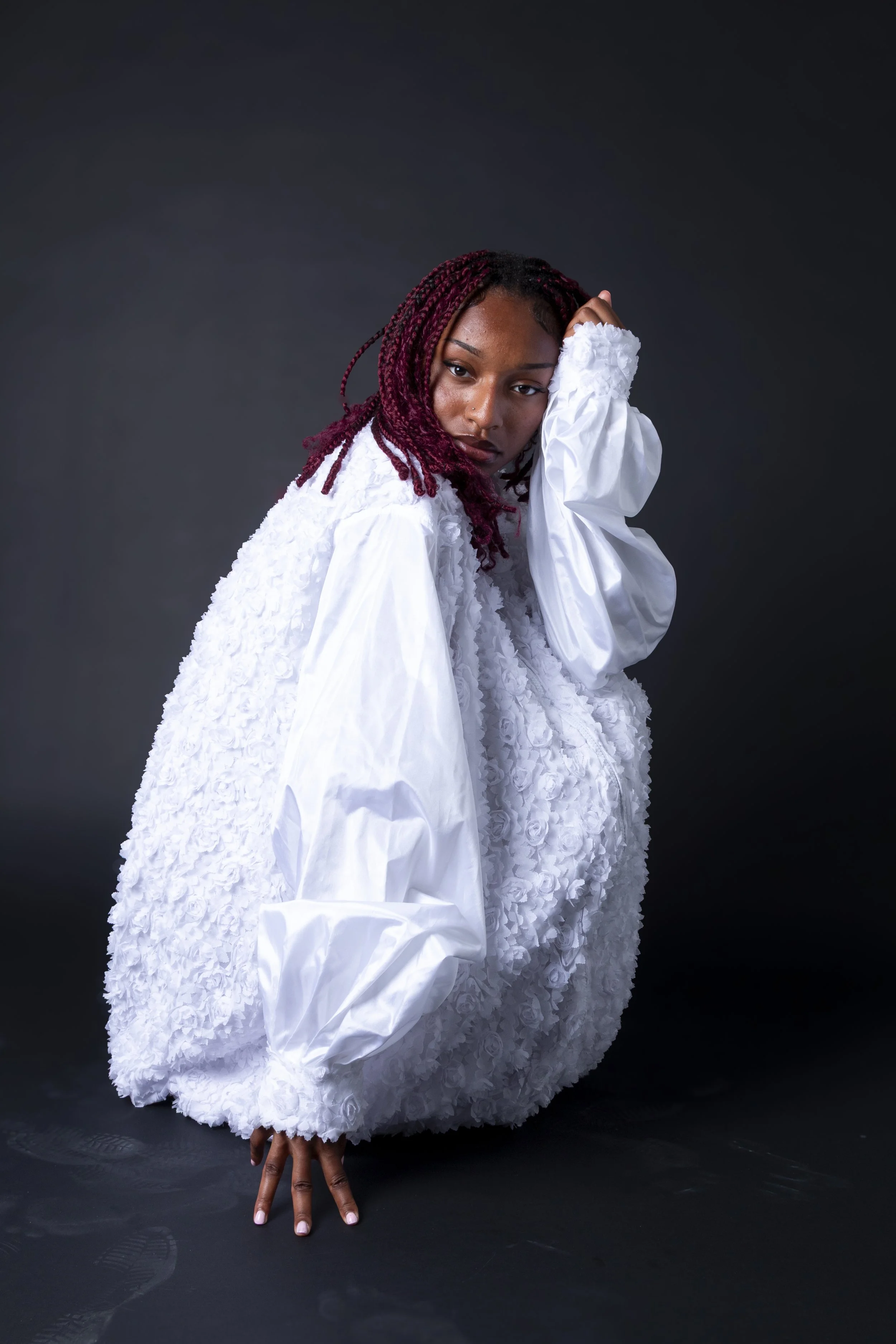 Person with braided red hair wearing a white textured dress, posing against a dark background, showing hand with fingers extended towards the bottom of the image.