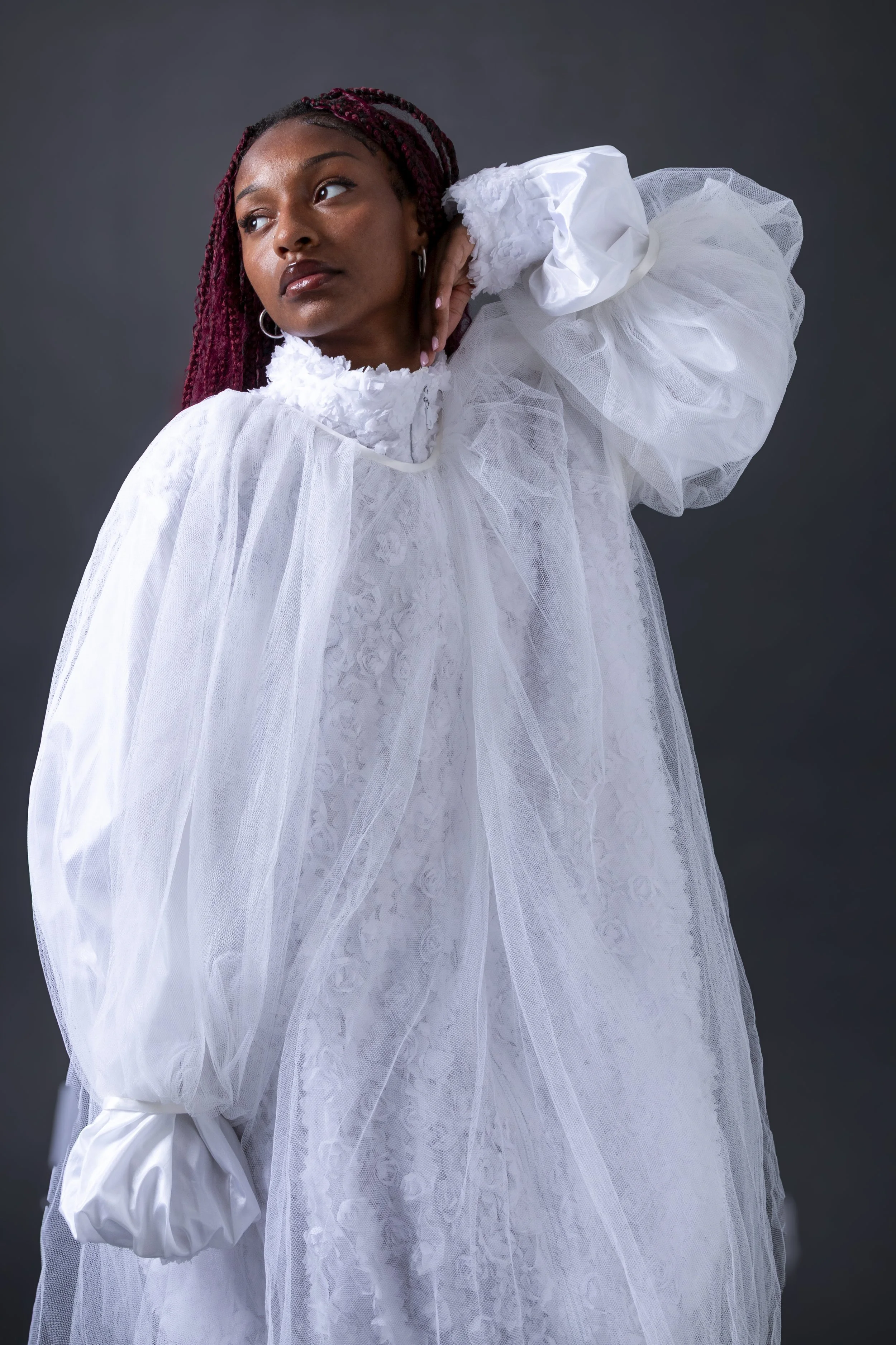 Portrait of a woman with braided hair wearing a white, ruffled, mesh dress, standing against a dark background.