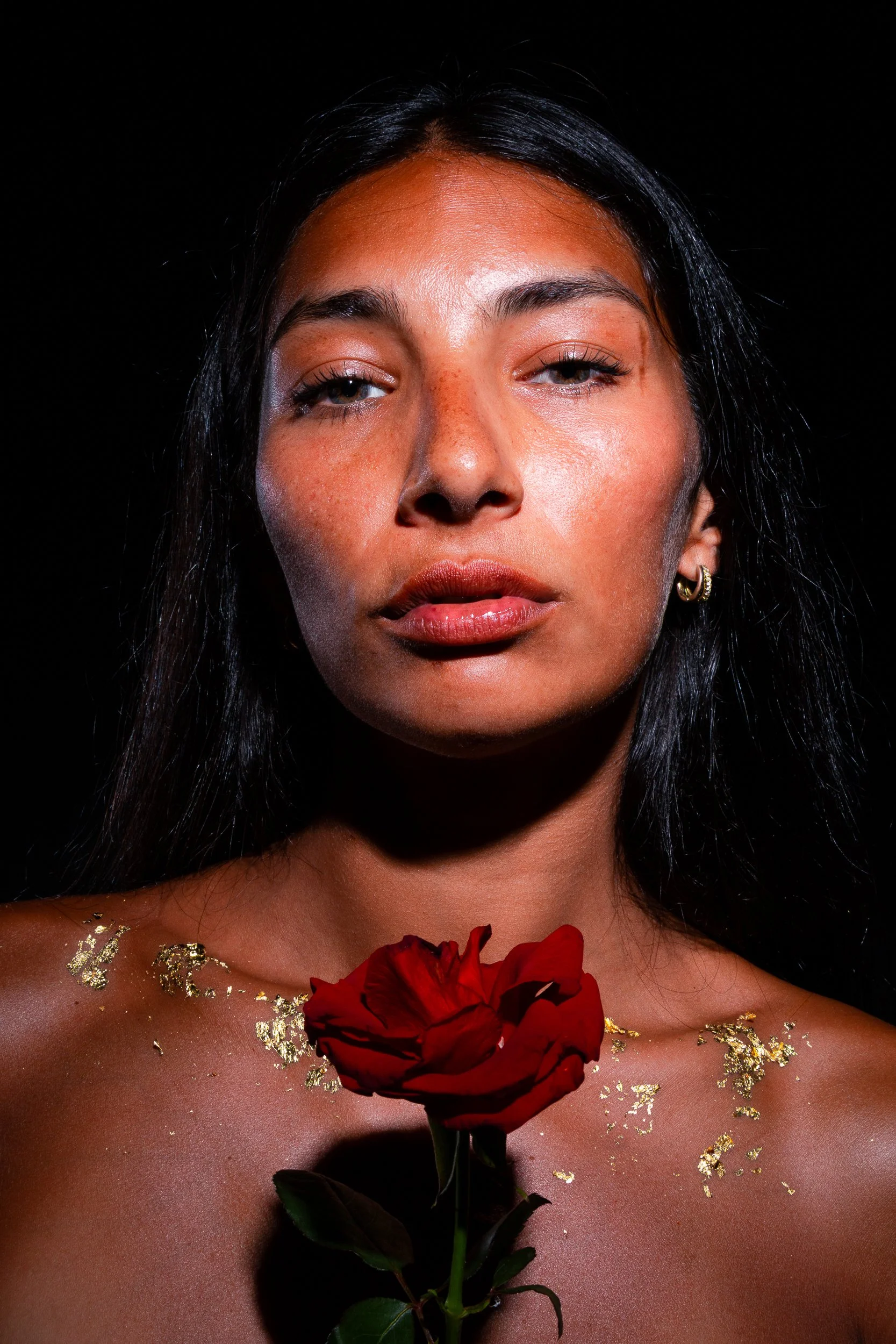 A woman with dark hair and earrings holding a red rose against her chest, with gold leaf accents on her skin, black background.