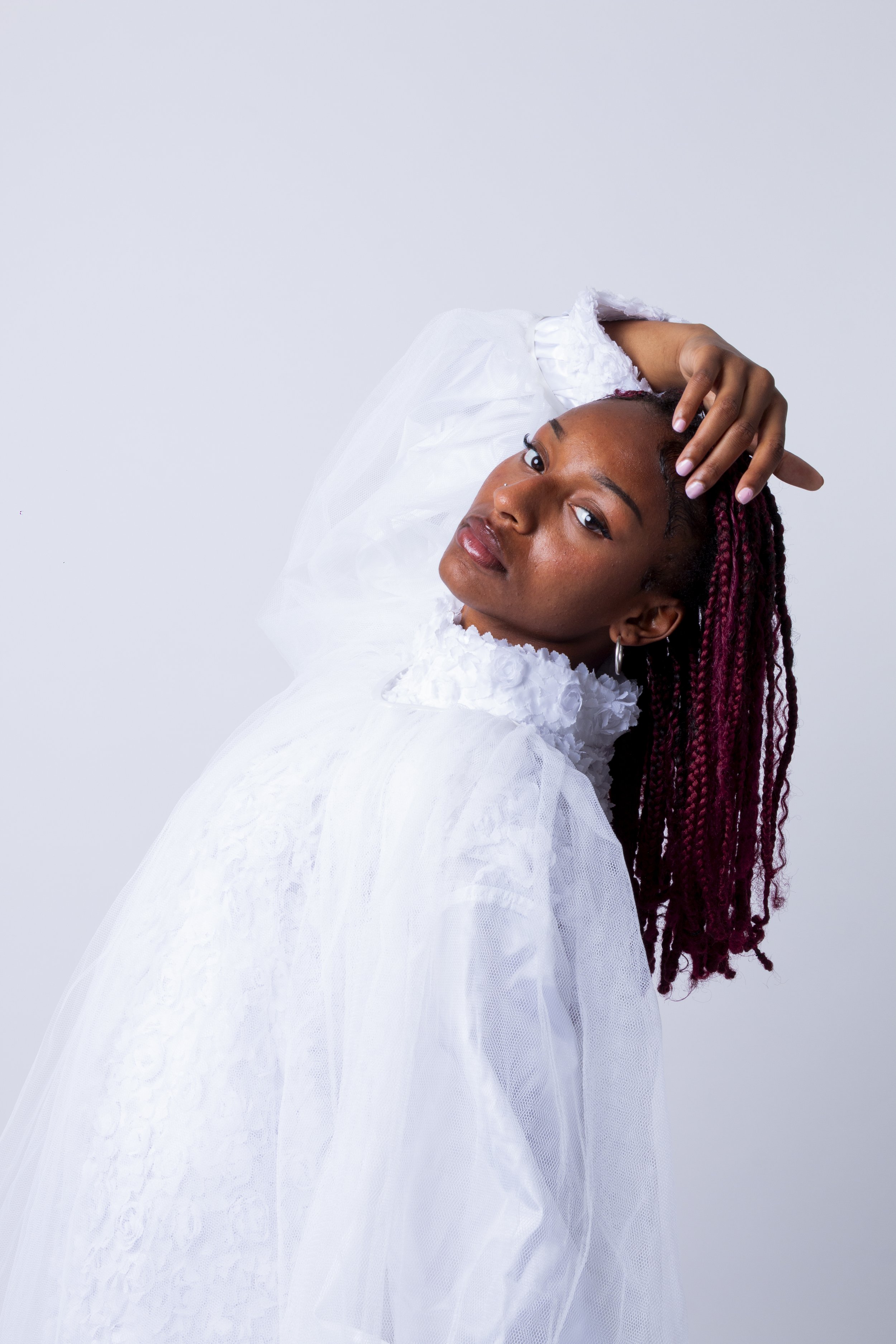 Portrait of a young Black woman with long reddish-brown braids, wearing a white dress with textured fabric, posed against a plain light background.