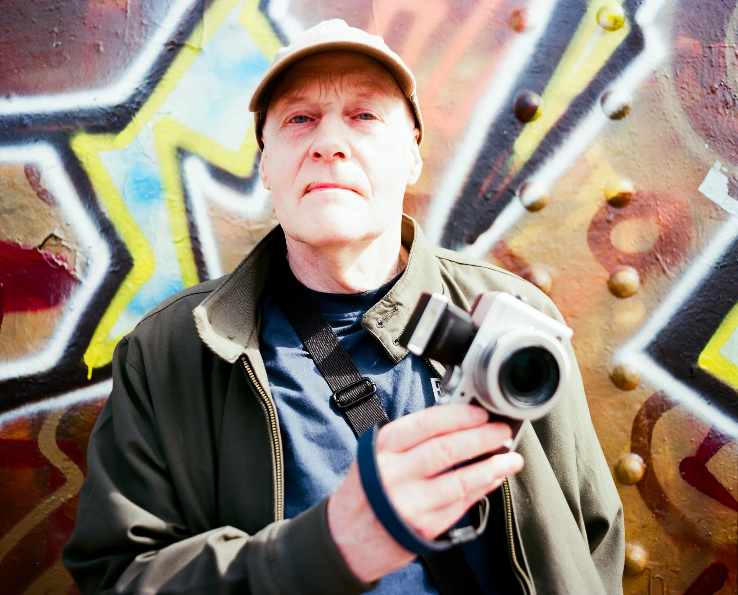 A man with light skin, wearing a beige cap and a green jacket, holding a camera in his right hand, stands in front of a colorful graffiti wall.