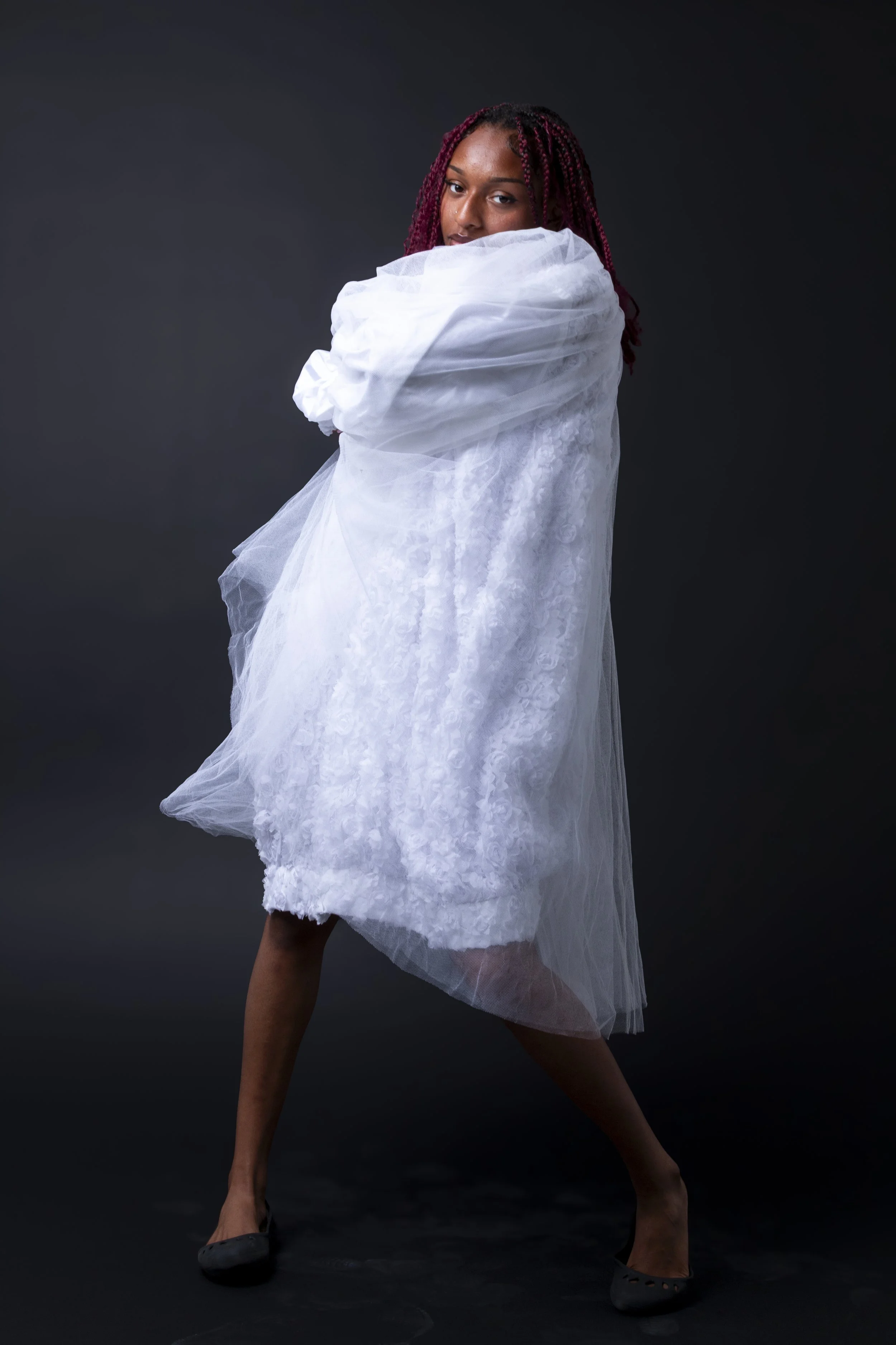 A woman with red braids posing against a dark background, wearing a white, textured, semi-transparent dress and black shoes.