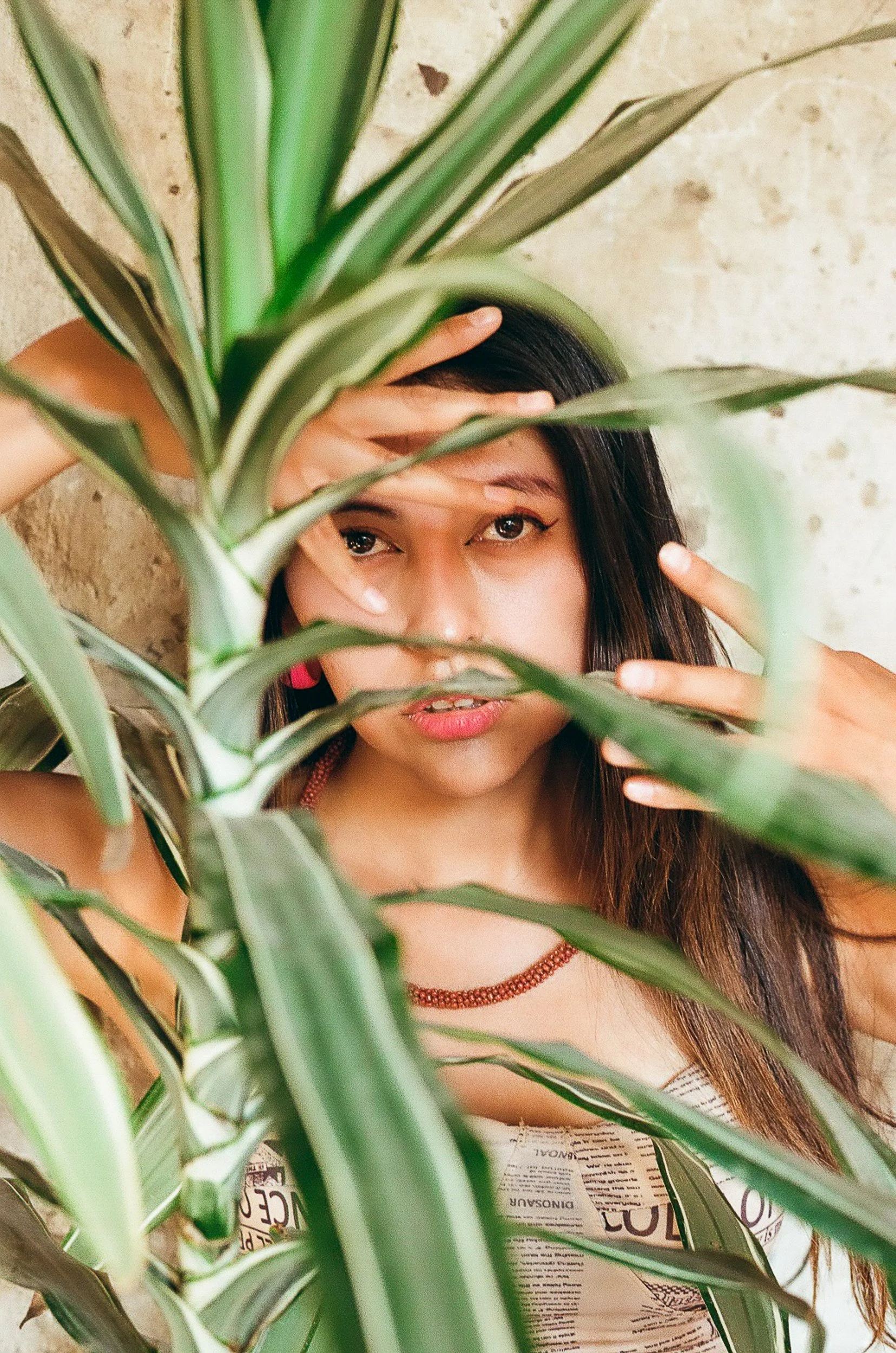 A woman with long dark hair and light skin is partially hidden behind a large green plant. She is touching her head with one hand and pointing with the other hand. The background is a beige textured wall. She is wearing a white shirt with printed tex