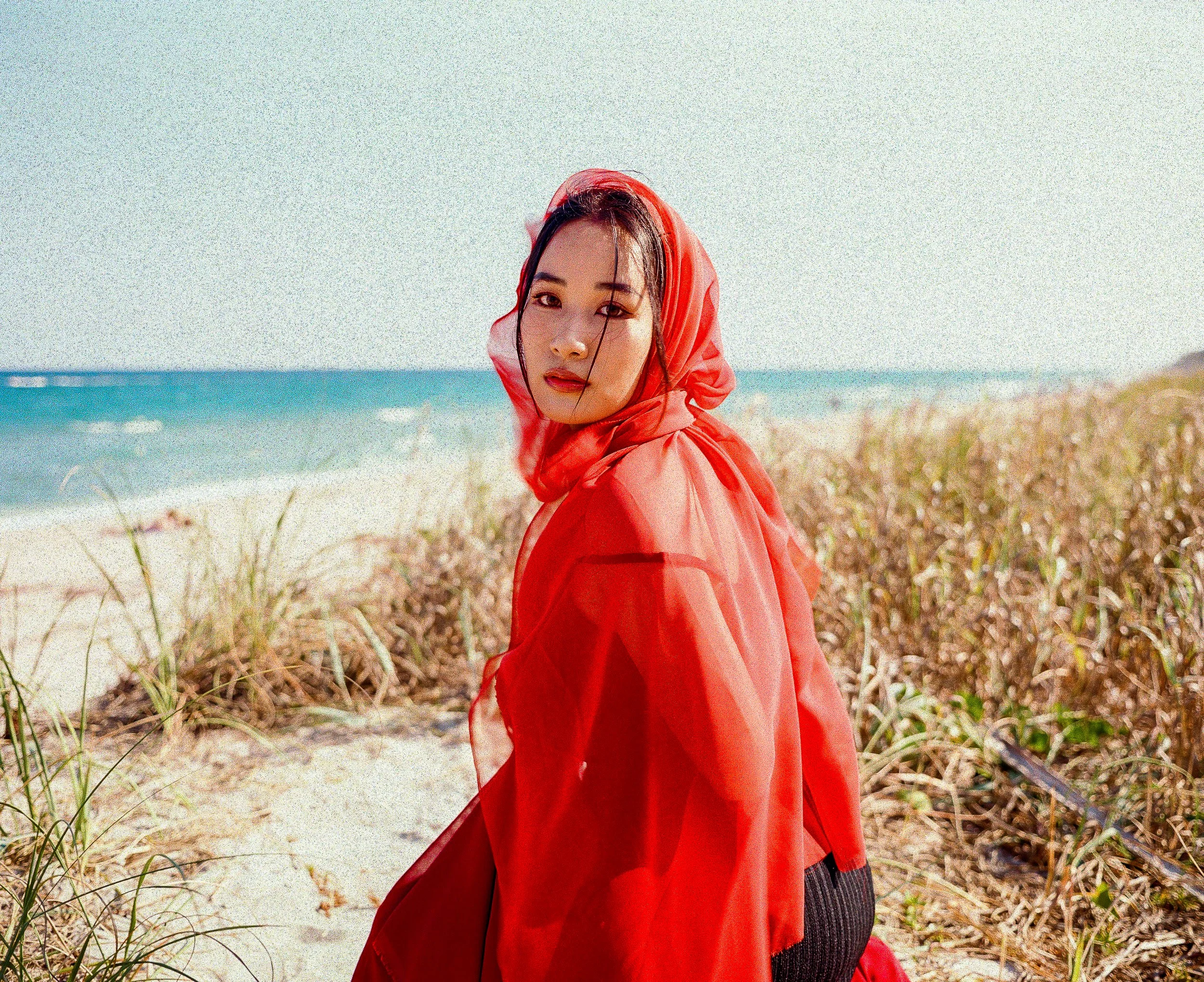 A woman in a red headscarf and matching red dress standing on a beach with sand dunes and greenery, and the ocean in the background.
