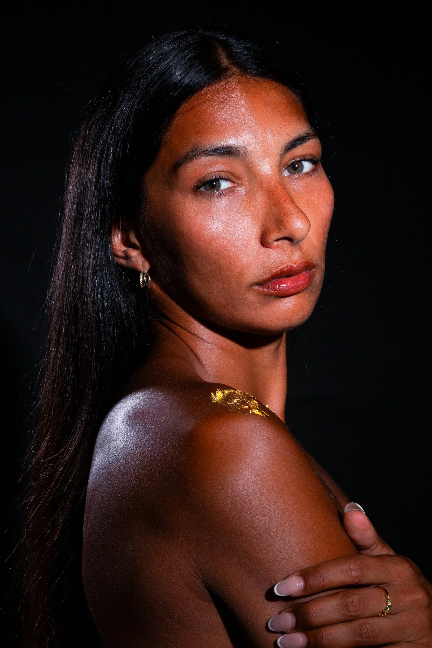 Close-up portrait of a woman with long dark hair, wearing gold earrings, with her arm crossed over her chest, against a black background.