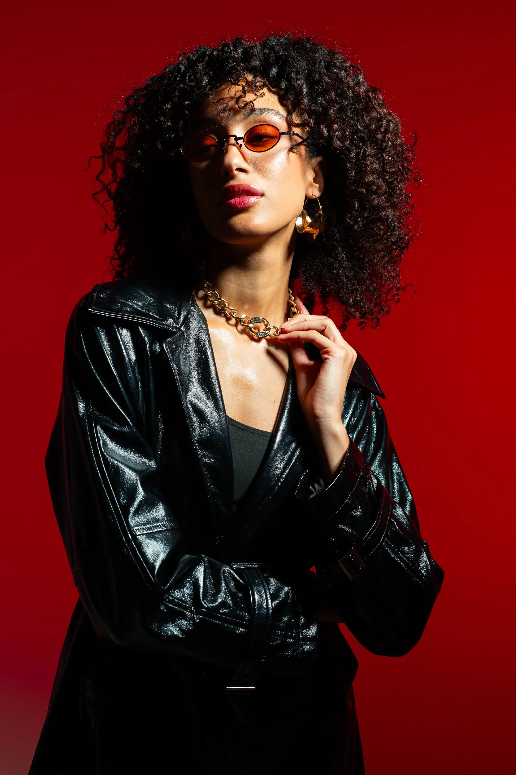 A woman with curly hair wearing a black leather jacket, orange-tinted sunglasses, bold red lipstick, gold hoop earrings, and a gold chain necklace against a red background.