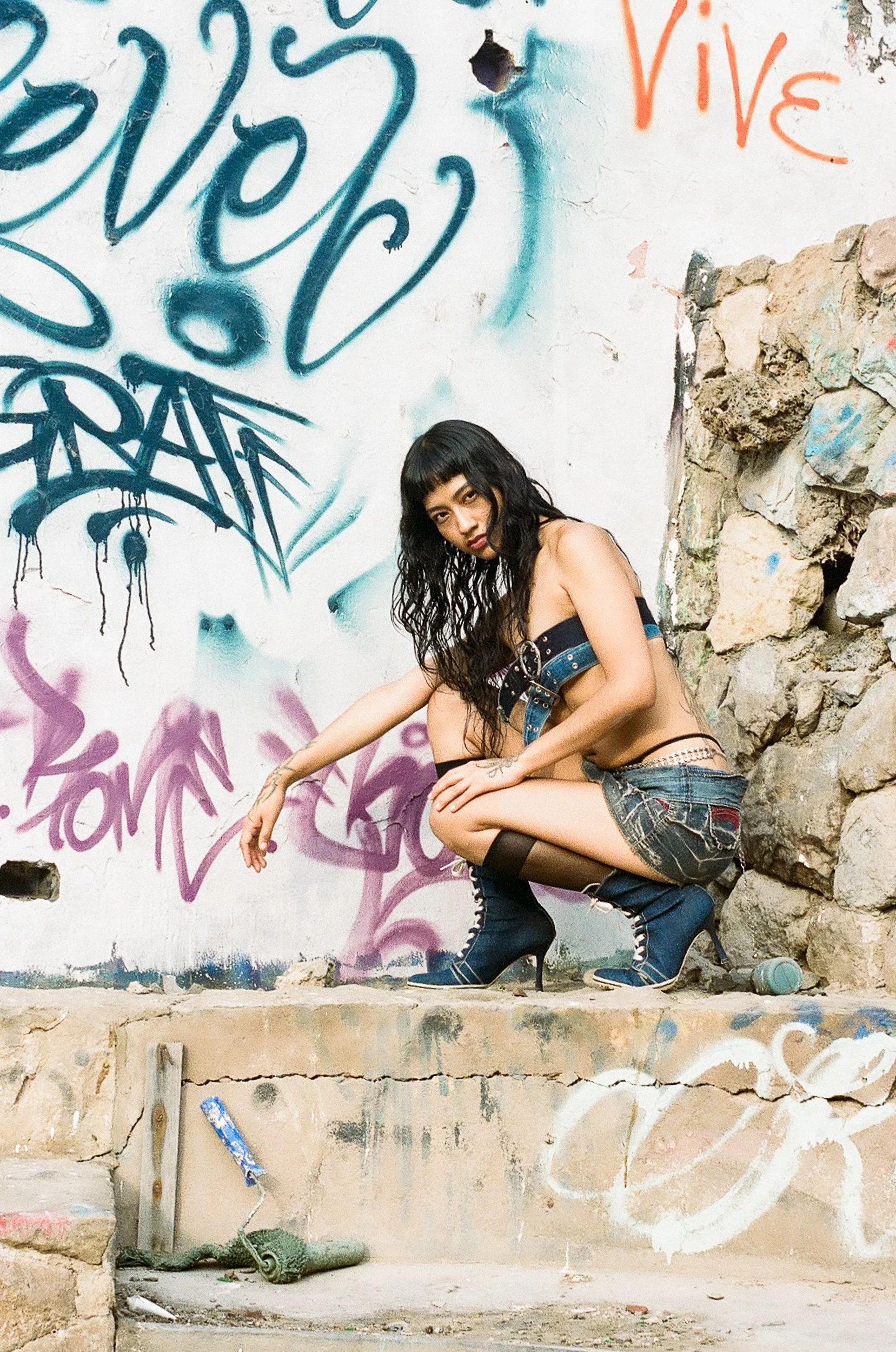 A woman with dark hair and bangs crouches against a graffiti-covered wall in an urban setting. She is wearing a black crop top, denim shorts, black stockings, and high-heeled boots. The wall behind her features various graffiti tags and phrases in bl