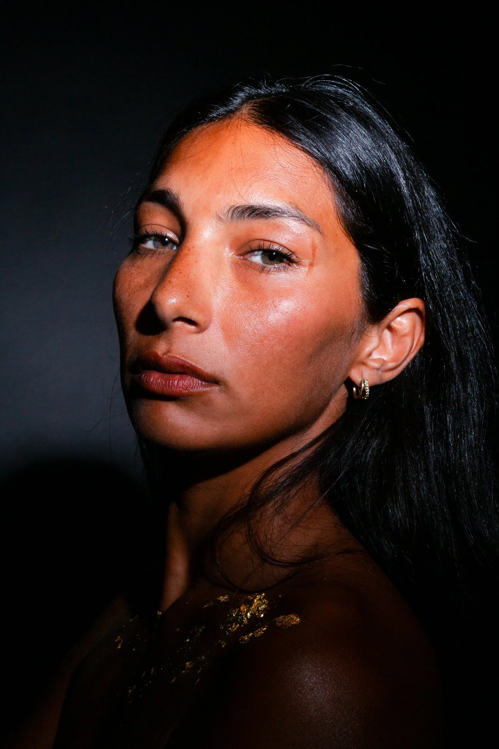 A close-up of a woman's face with dark hair, light skin, light eye makeup, and gold earrings, against a black background.