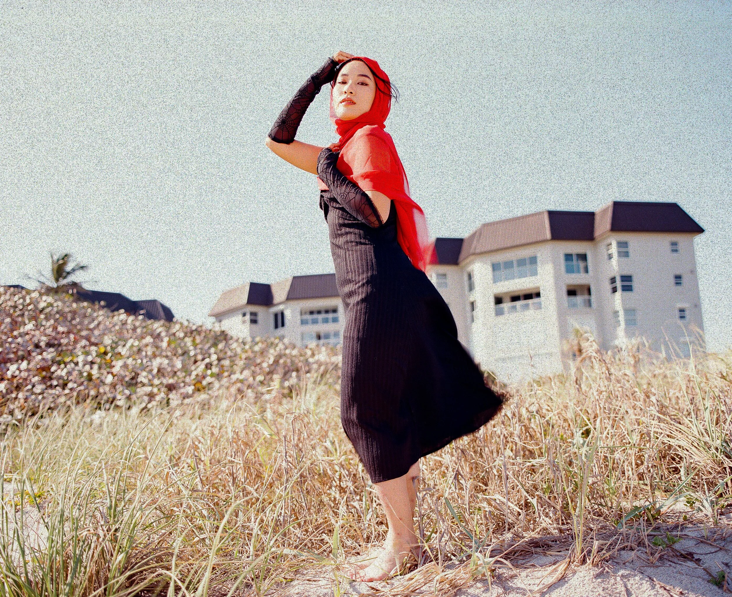 A woman with a red headscarf and black dress standing in a field of tall grass, with modern white buildings in the background.