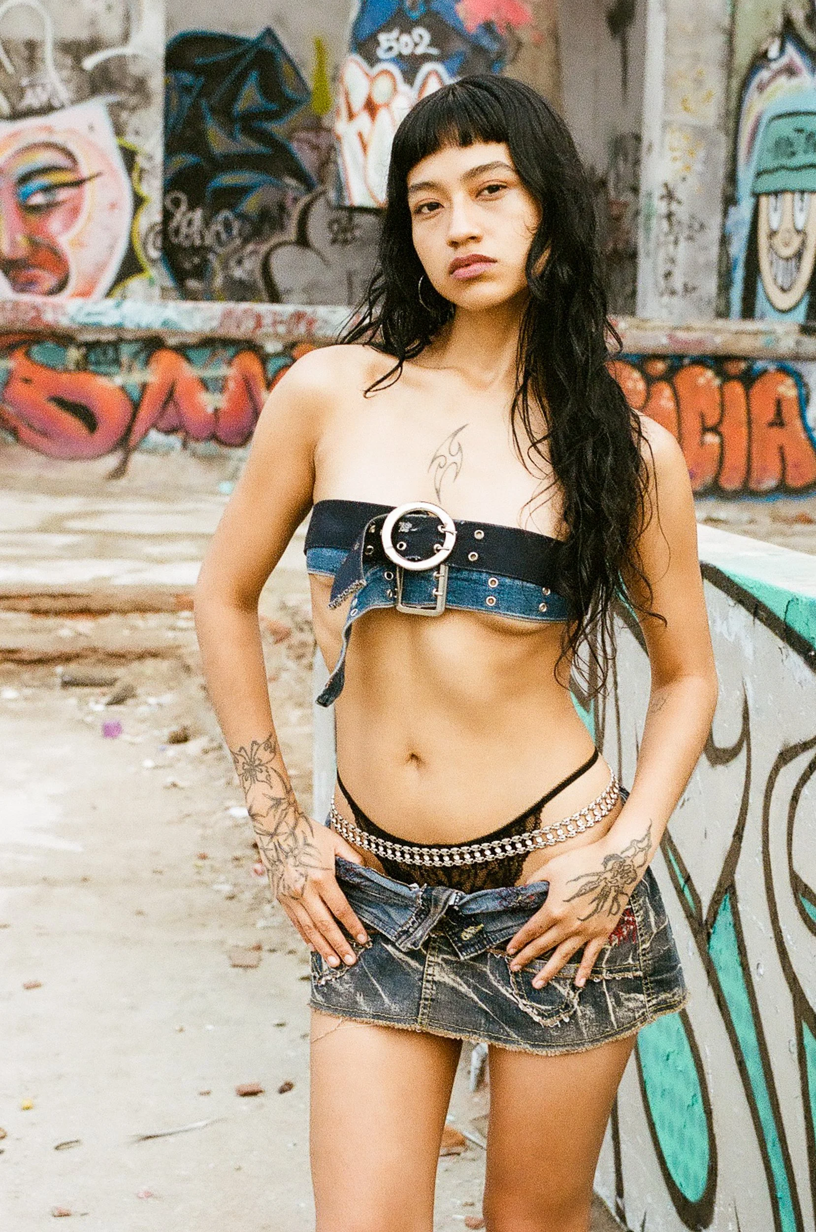 A woman with long black hair, tattoos, and hoop earrings stands against a graffiti-covered wall, wearing a black strapless top with a silver buckle, ripped denim shorts, and chain jewelry.