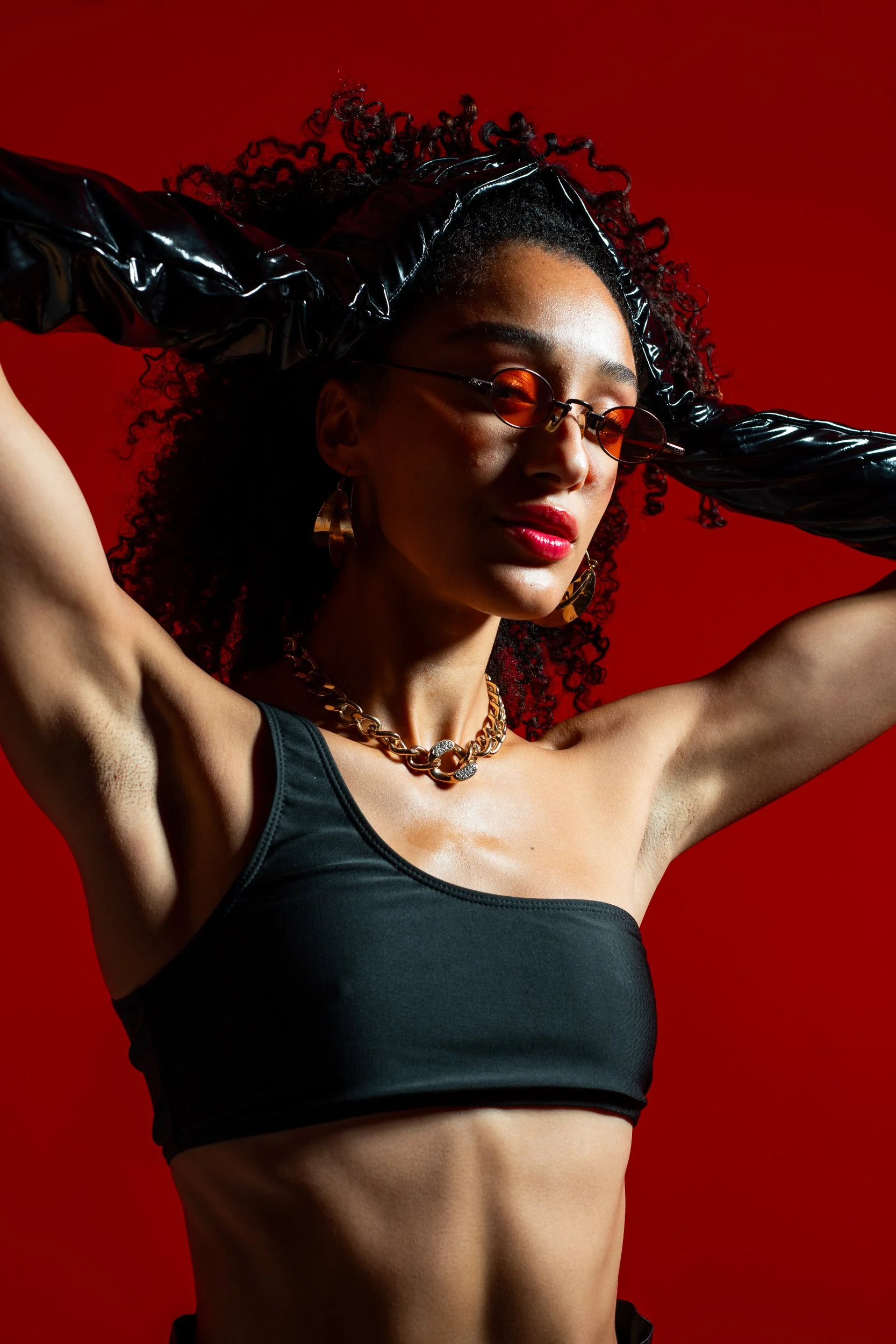 A woman with curly dark hair, wearing orange-tinted sunglasses, gold hoop earrings, a chunky gold chain necklace, and a black sports bra, poses against a red background with her arms raised.