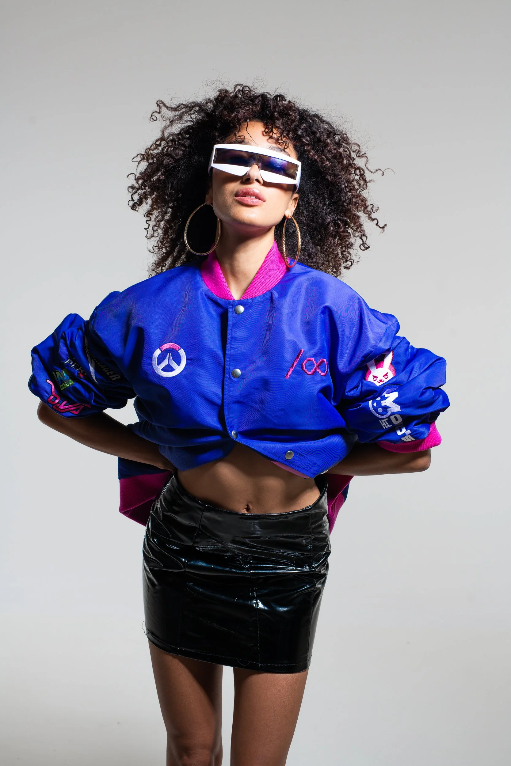 A woman with curly hair wearing large sunglasses, a blue bomber jacket with various patches, and a black latex skirt.