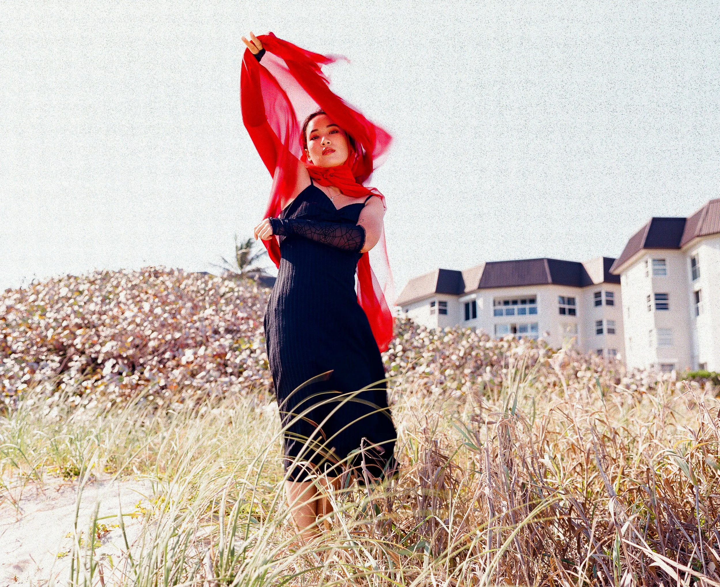 A woman wearing a black dress and red scarf stands in a grassy field, holding a sheer red fabric above her head, with residential buildings in the background.