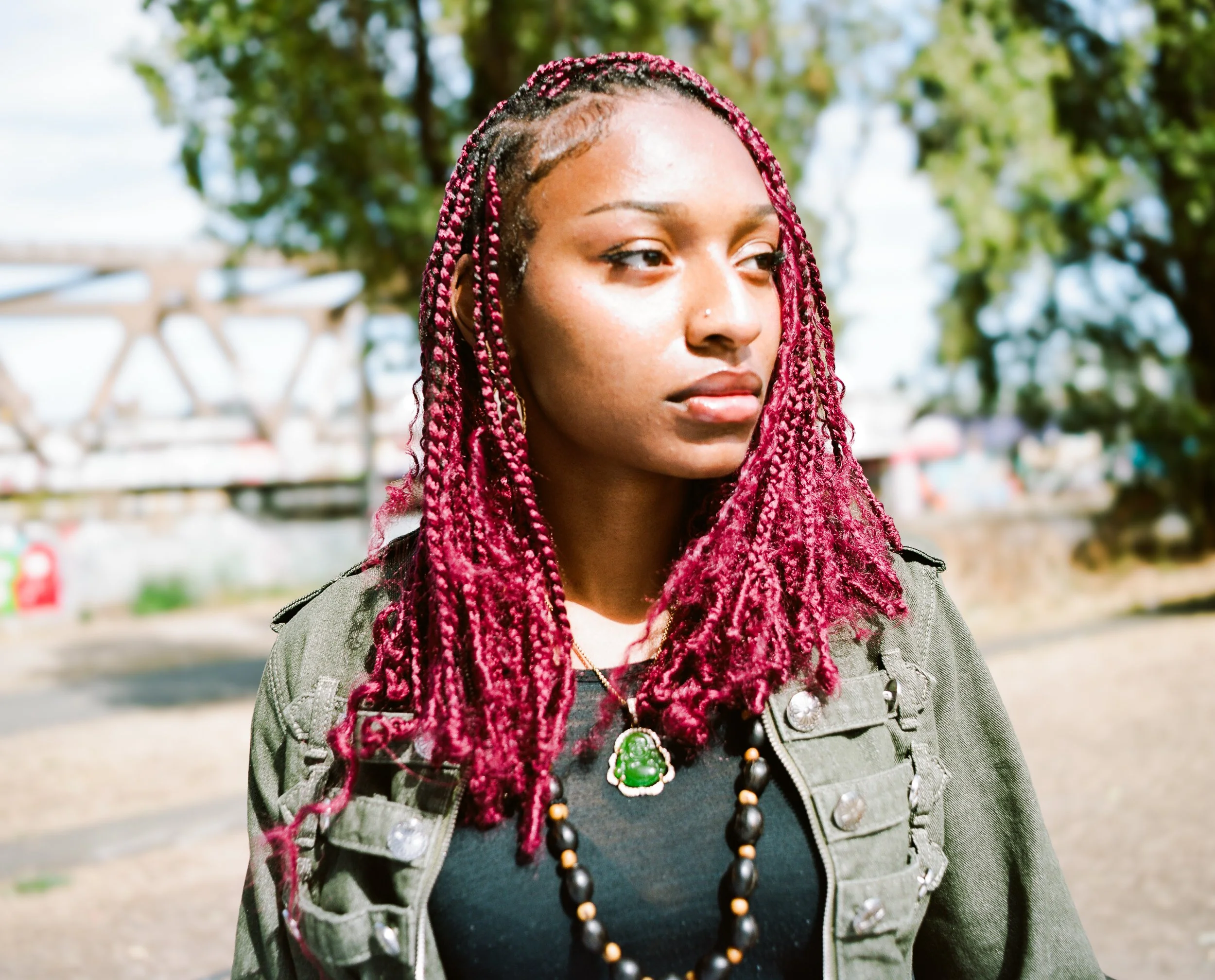 A woman with red braided hair, wearing a gray jacket and a necklace with a large green pendant, standing outdoors near trees.