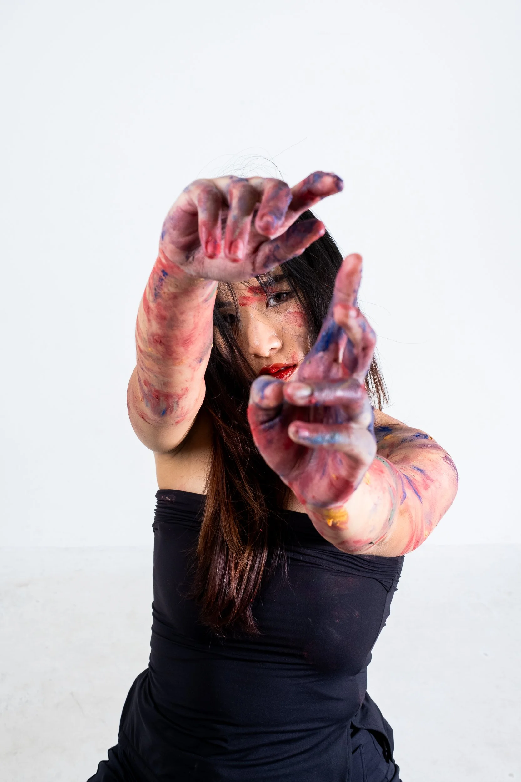 Woman with long brown hair wearing a black top, covered in colorful paint, reaching out with her right hand towards the camera against a plain white background.